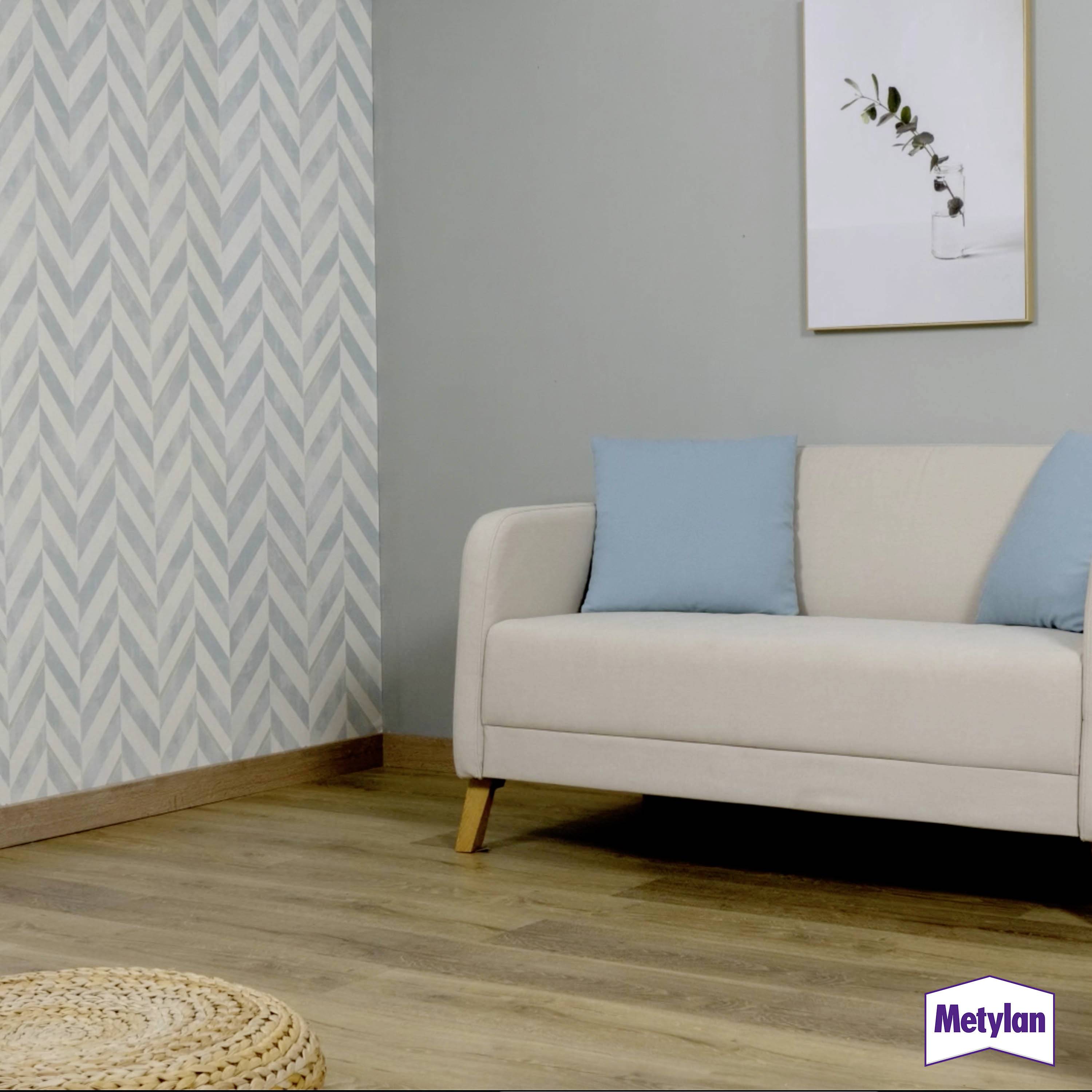 Un salon avec un canapé beige, des coussins bleus, un sol en bois clair, et un papier peint à motifs bleu-gris. Une photo encadrée de plante est accrochée au mur.