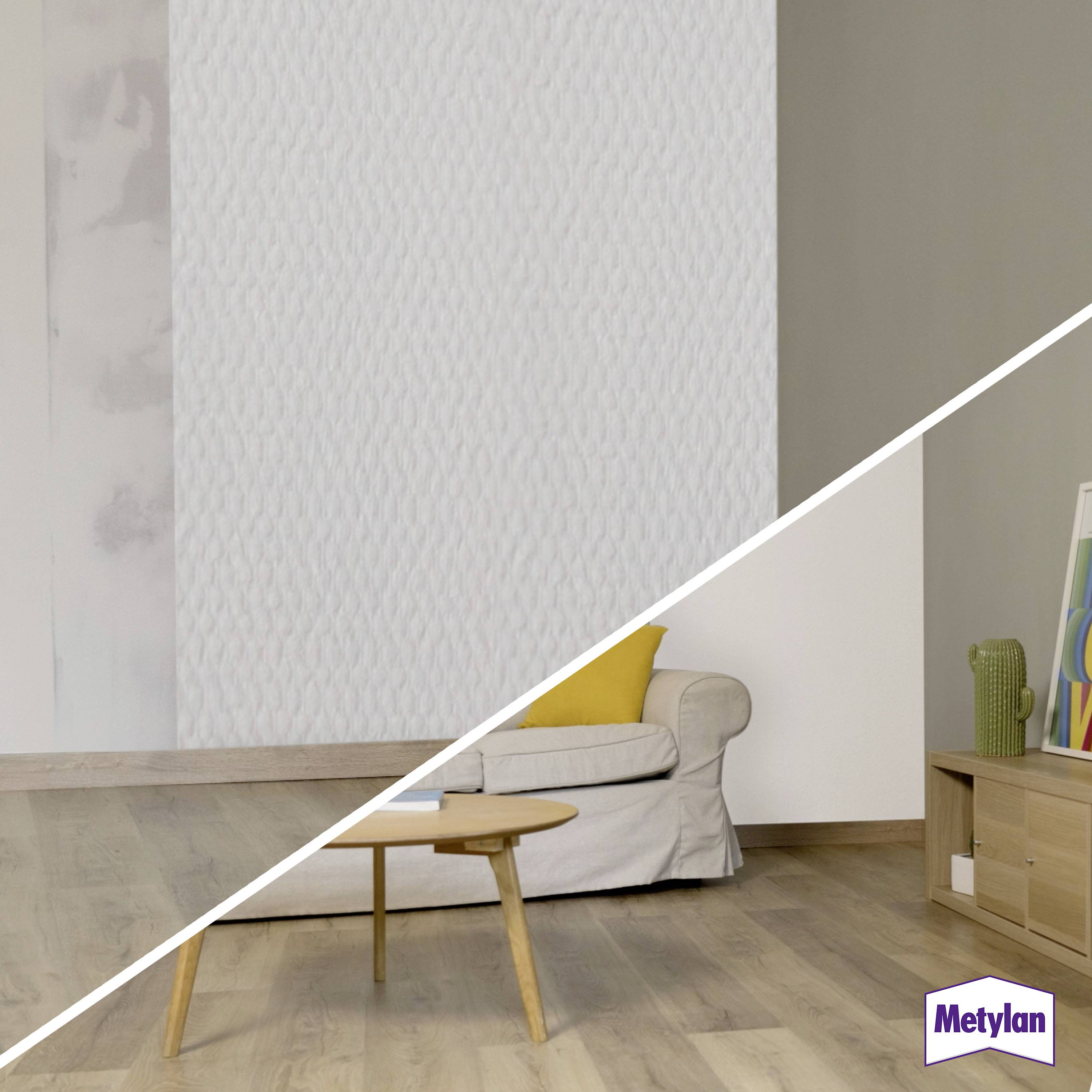 Un salon avec un mur partiellement tapissé et un canapé beige sur un sol en bois ; le papier peint présente un motif léger et texturé.
