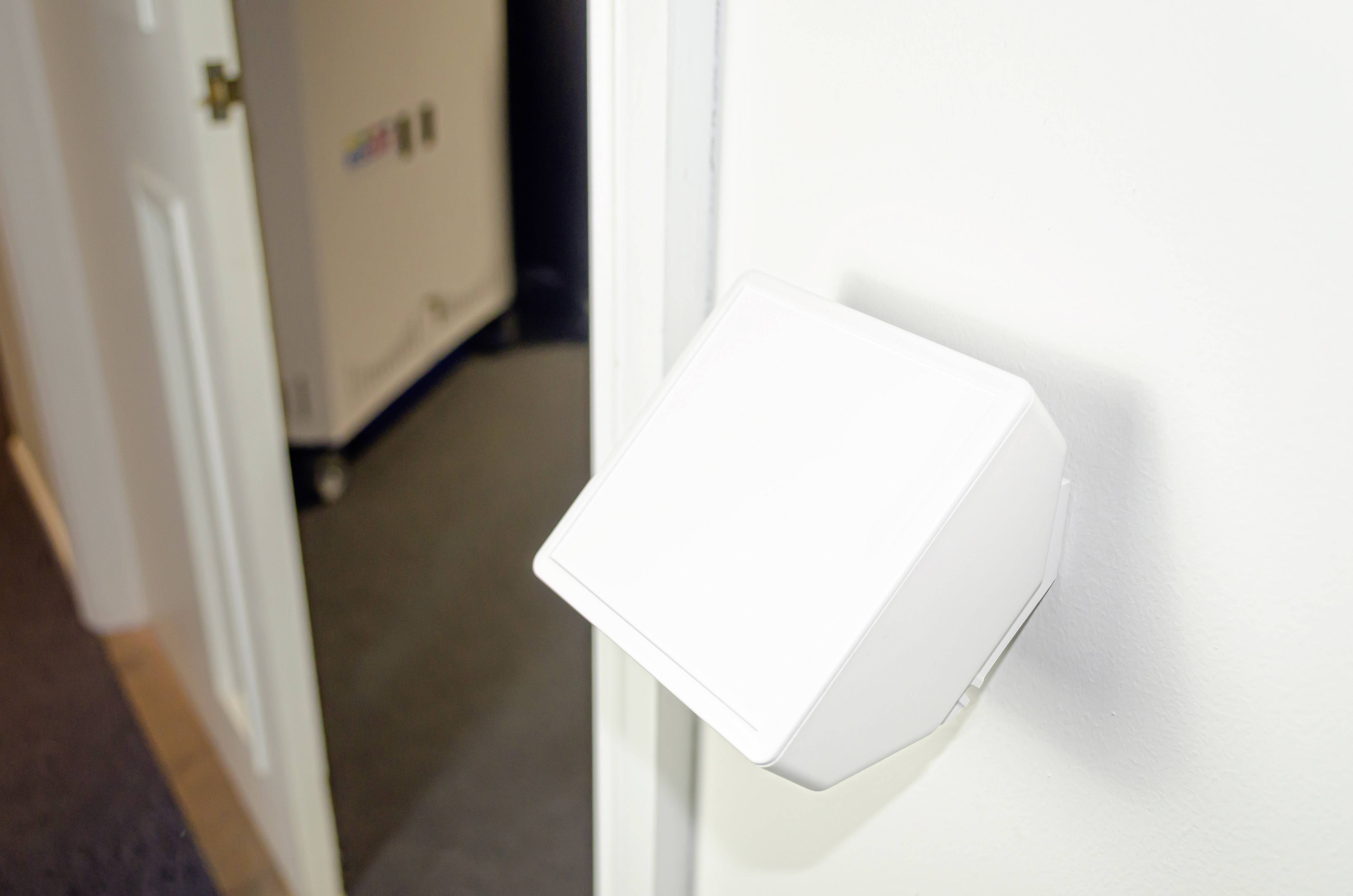 Un capteur blanc et carré est suspendu à un mur dans une pièce dont la porte est ouverte.
