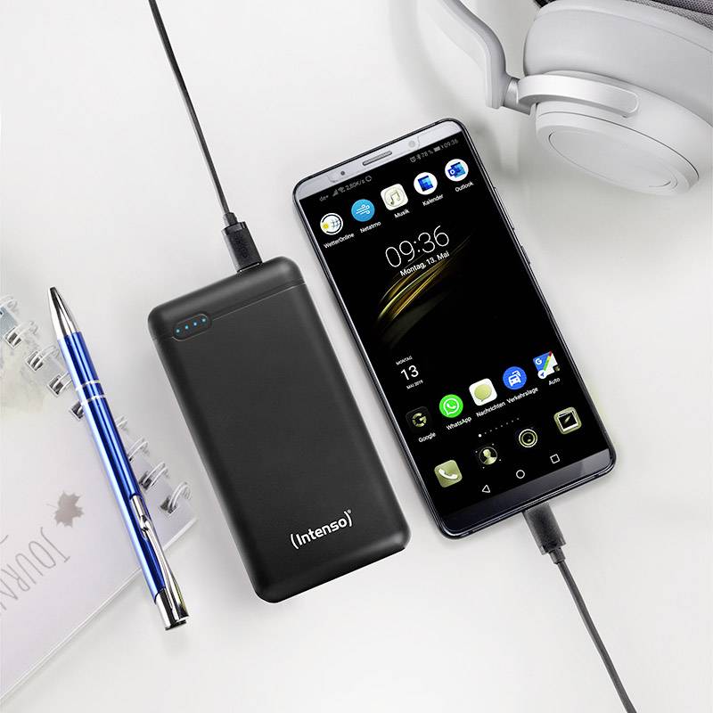 Un smartphone et une batterie externe sont chargés côte à côte. Un carnet de notes et un stylo à bille sont posés à proximité. Des écouteurs sont visibles dans le coin.
