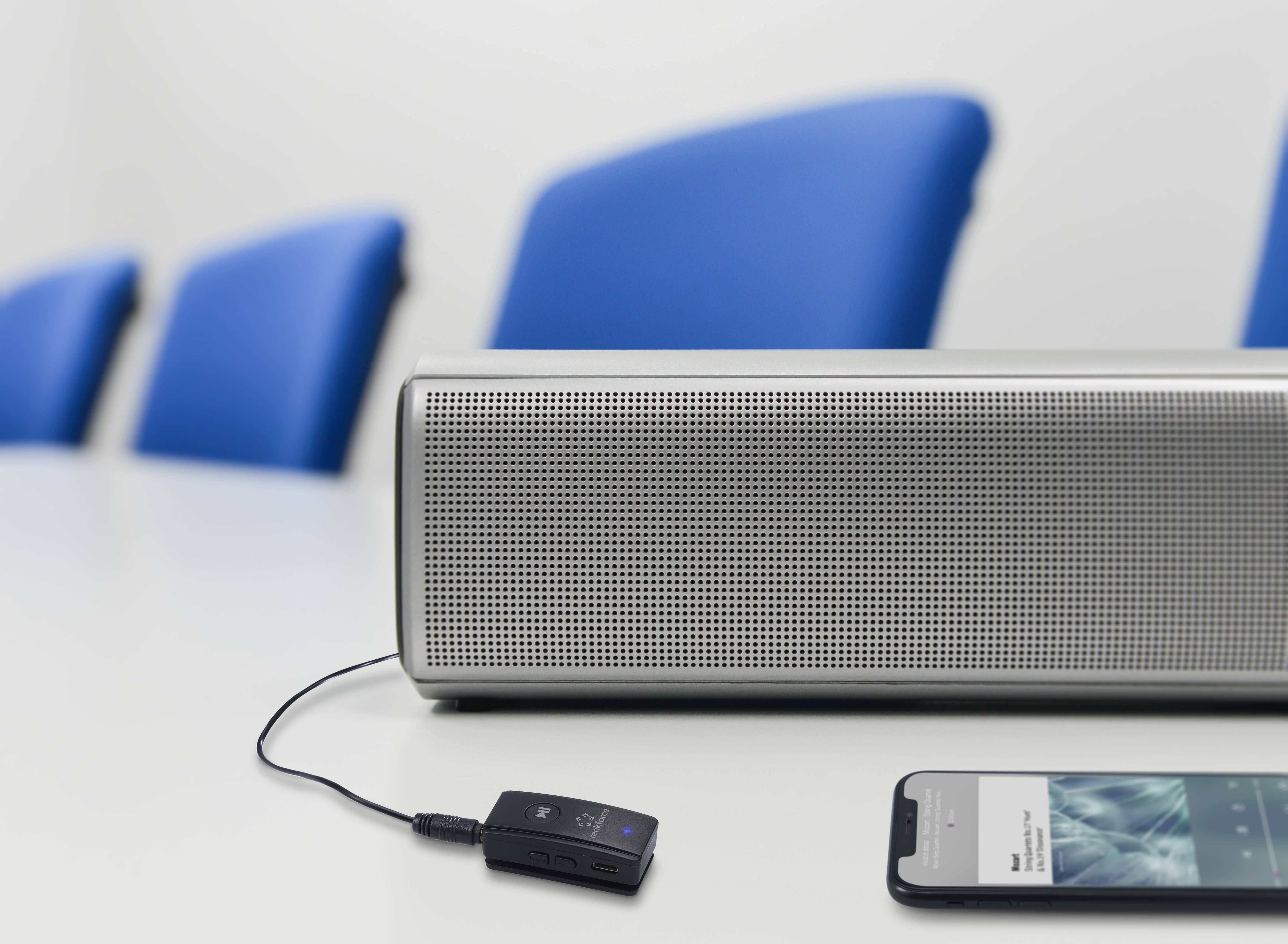 Un smartphone est connecté à un haut-parleur par un câble sur une table blanche. Des chaises bleues sont visibles en arrière-plan.