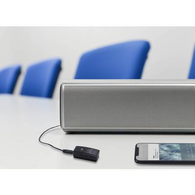 Un smartphone est connecté à un haut-parleur par un câble sur une table blanche. Des chaises bleues sont visibles en arrière-plan.