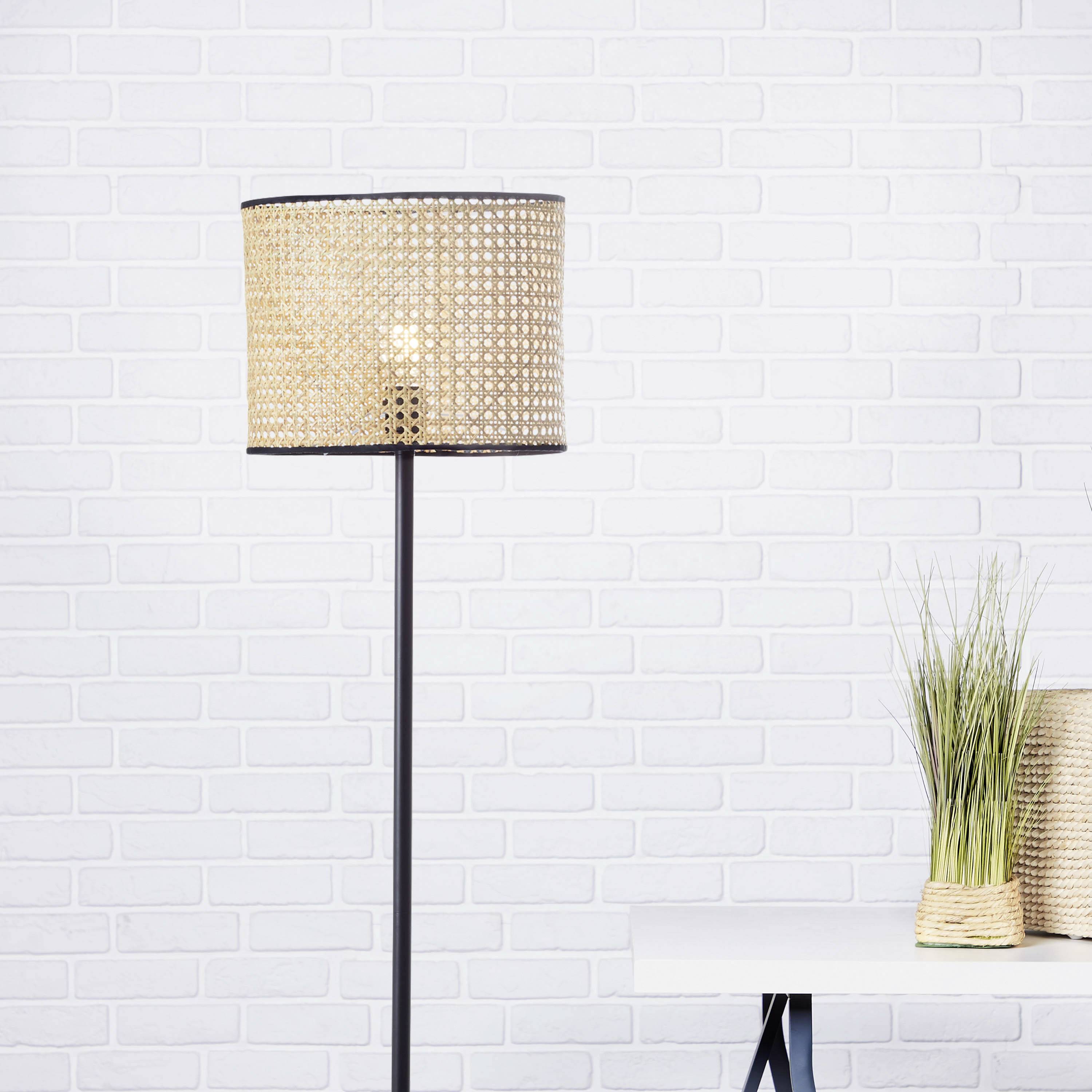 Lampadaire avec abat-jour tressé à côté d'une table avec une plante, devant un mur de briques blanches.