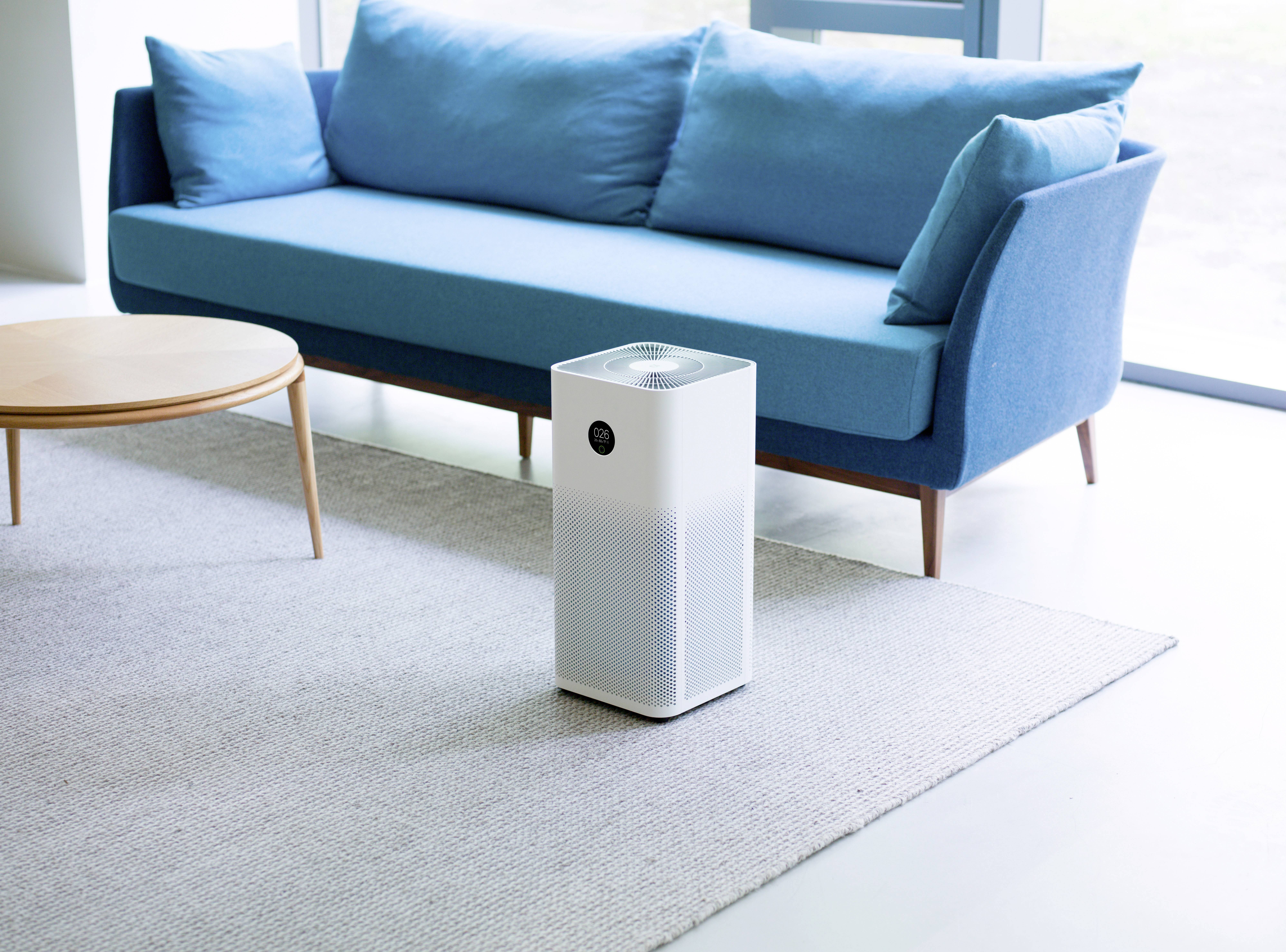 Un purificateur d'air blanc est placé sur un tapis devant un canapé bleu et une table ronde en bois dans une pièce au design moderne.