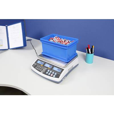 Une boîte bleue, remplie de tubes rouges et blancs, est posée sur une balance numérique sur un bureau. L'affichage indique '36,5 kg'.