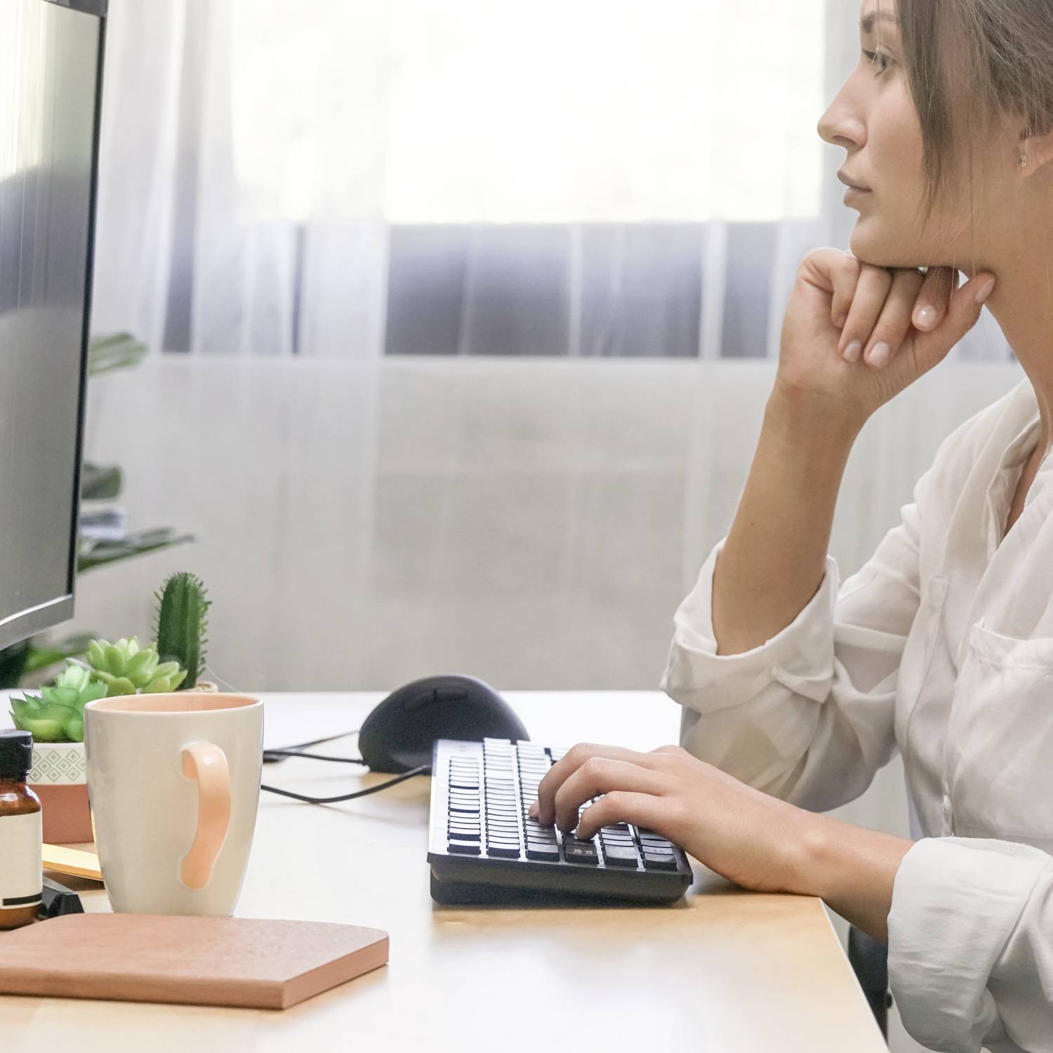 Une personne se concentre sur un écran d'ordinateur. Sur le bureau se trouvent une tasse de café et des plantes, la pièce est lumineuse.