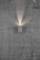 Un mur en béton gris avec une lampe murale cylindrique moderne qui projette sa lumière vers le haut contre le mur, créant un cône lumineux.