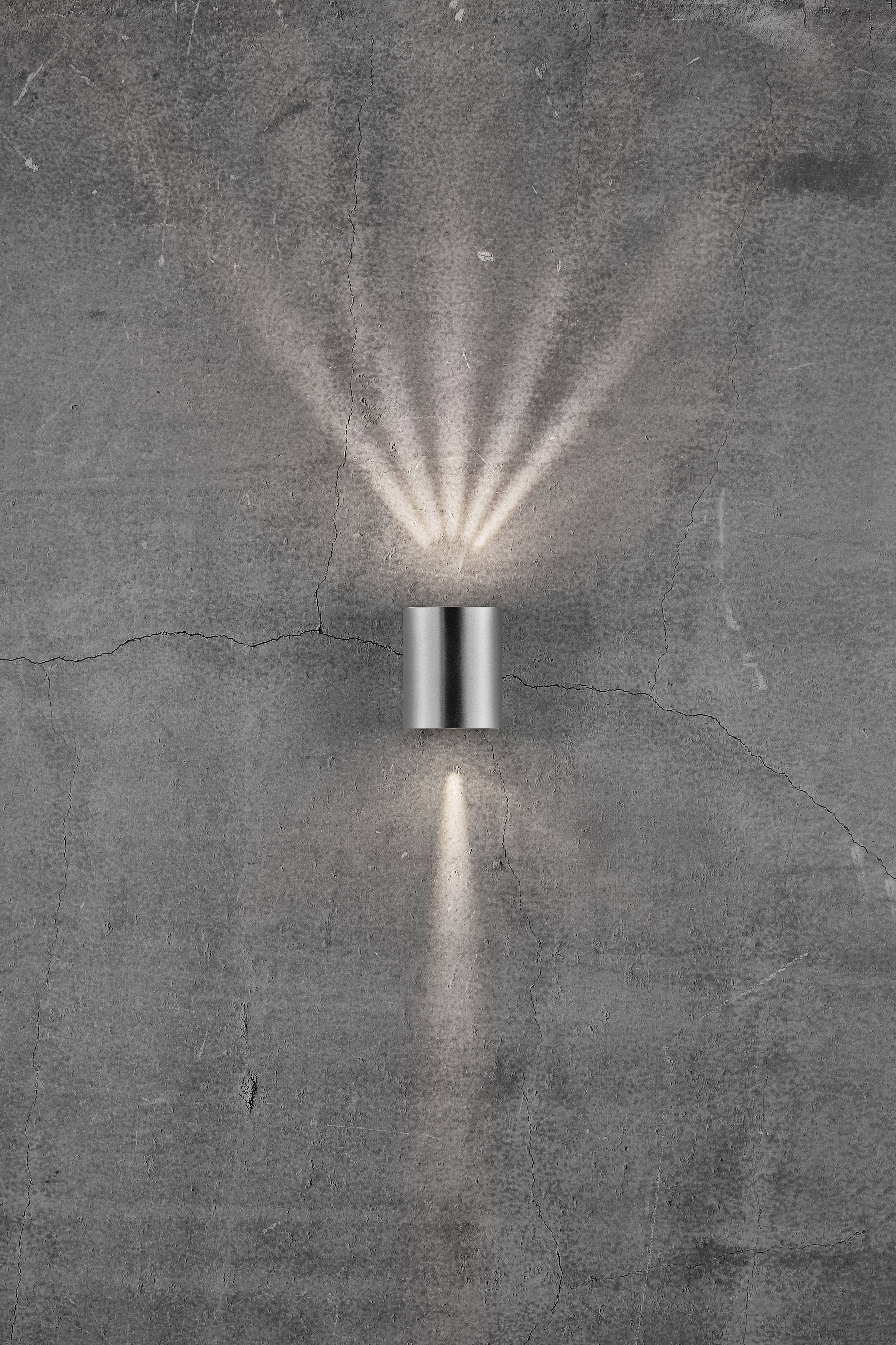 Lampe murale moderne en argent fixée à un mur en béton gris et fissuré. La lumière rayonne selon un motif conique vers le haut et vers le bas.