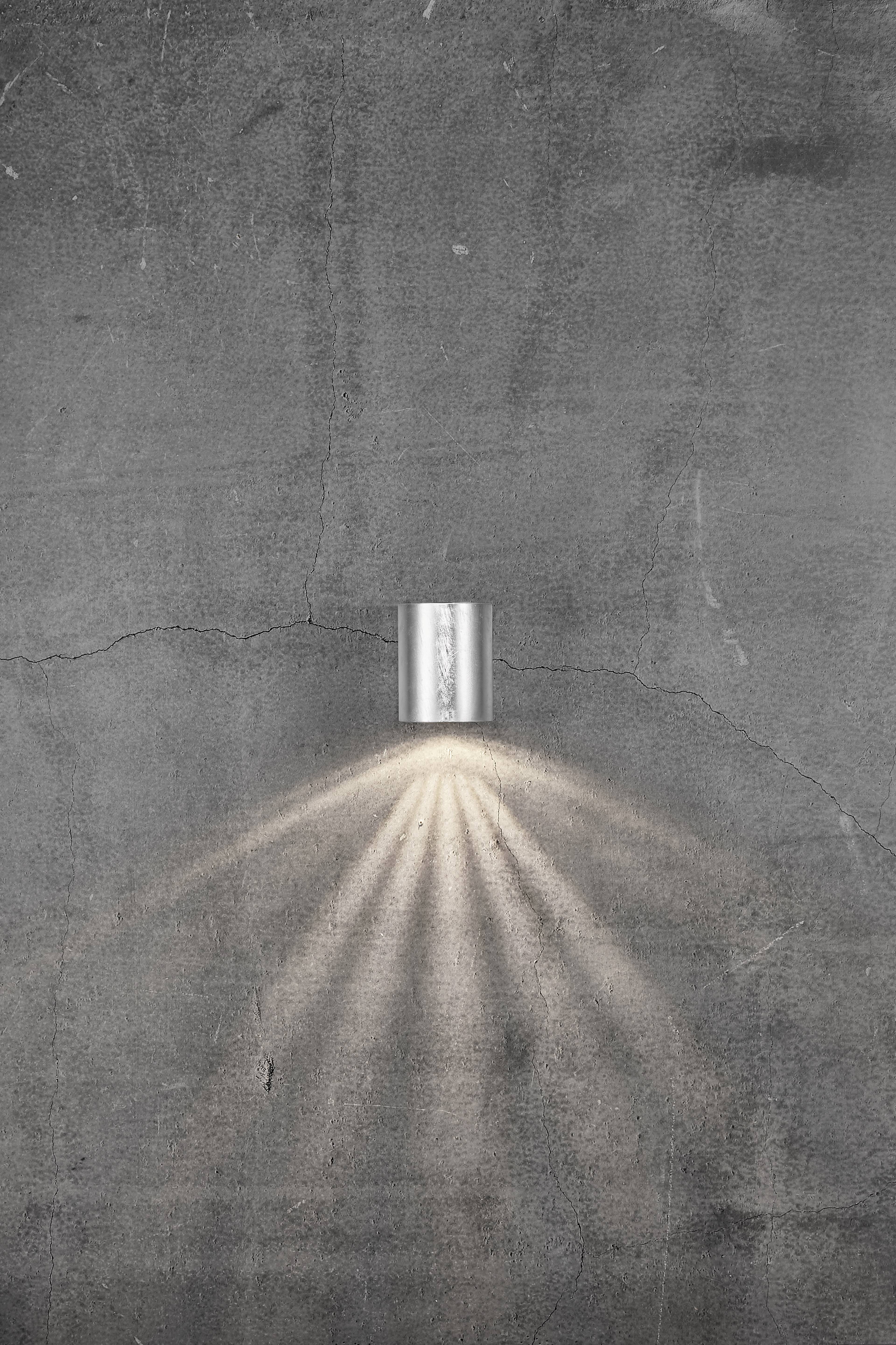 Un petit applique moderne en métal projette une lumière douce sur un mur de béton rugueux et gris. Les rayons lumineux se propagent en éventail.