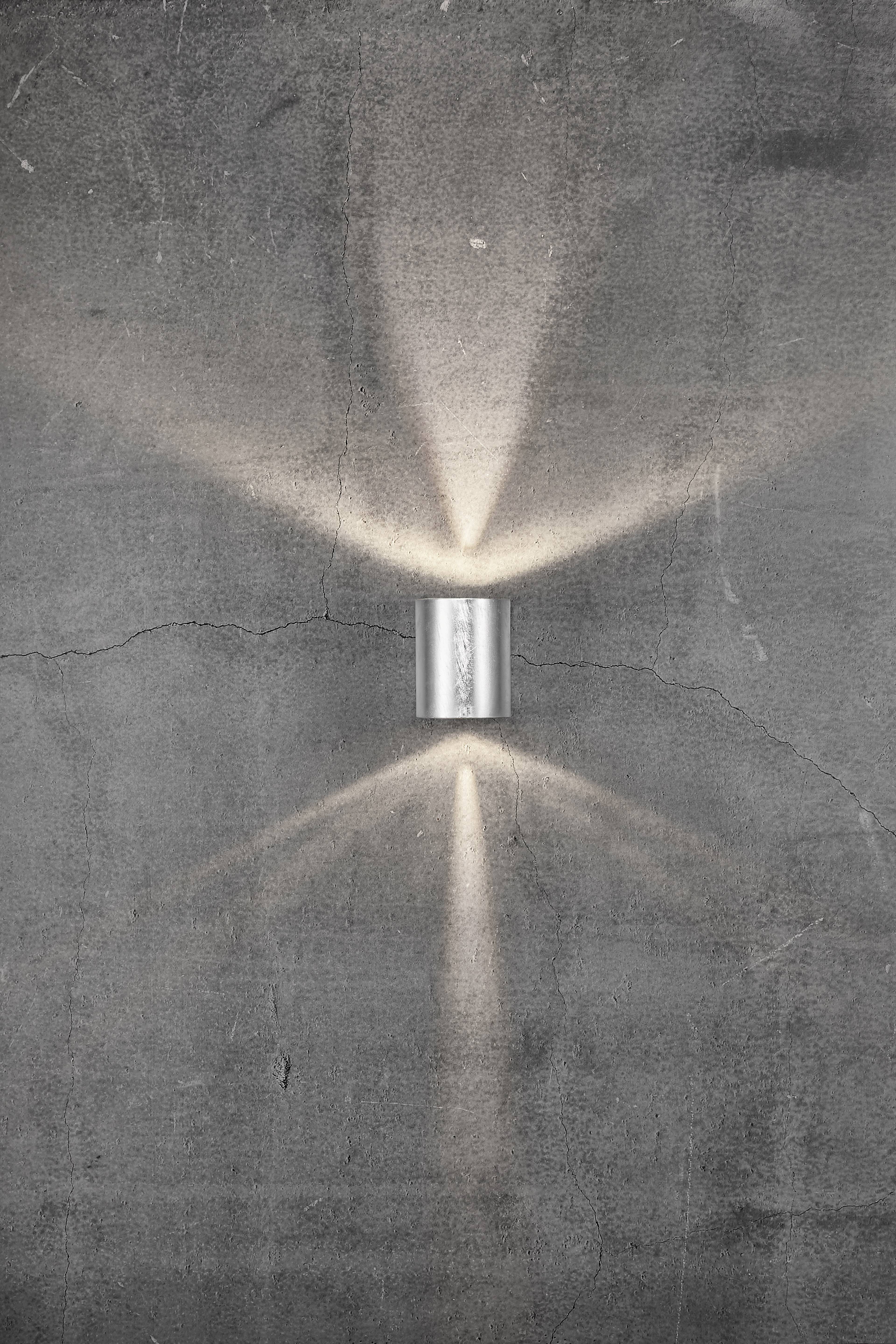 Un applique murale moderne en métal qui diffuse des rayons lumineux vers le haut et vers le bas sur un mur rugueux gris, créant un motif élégant.