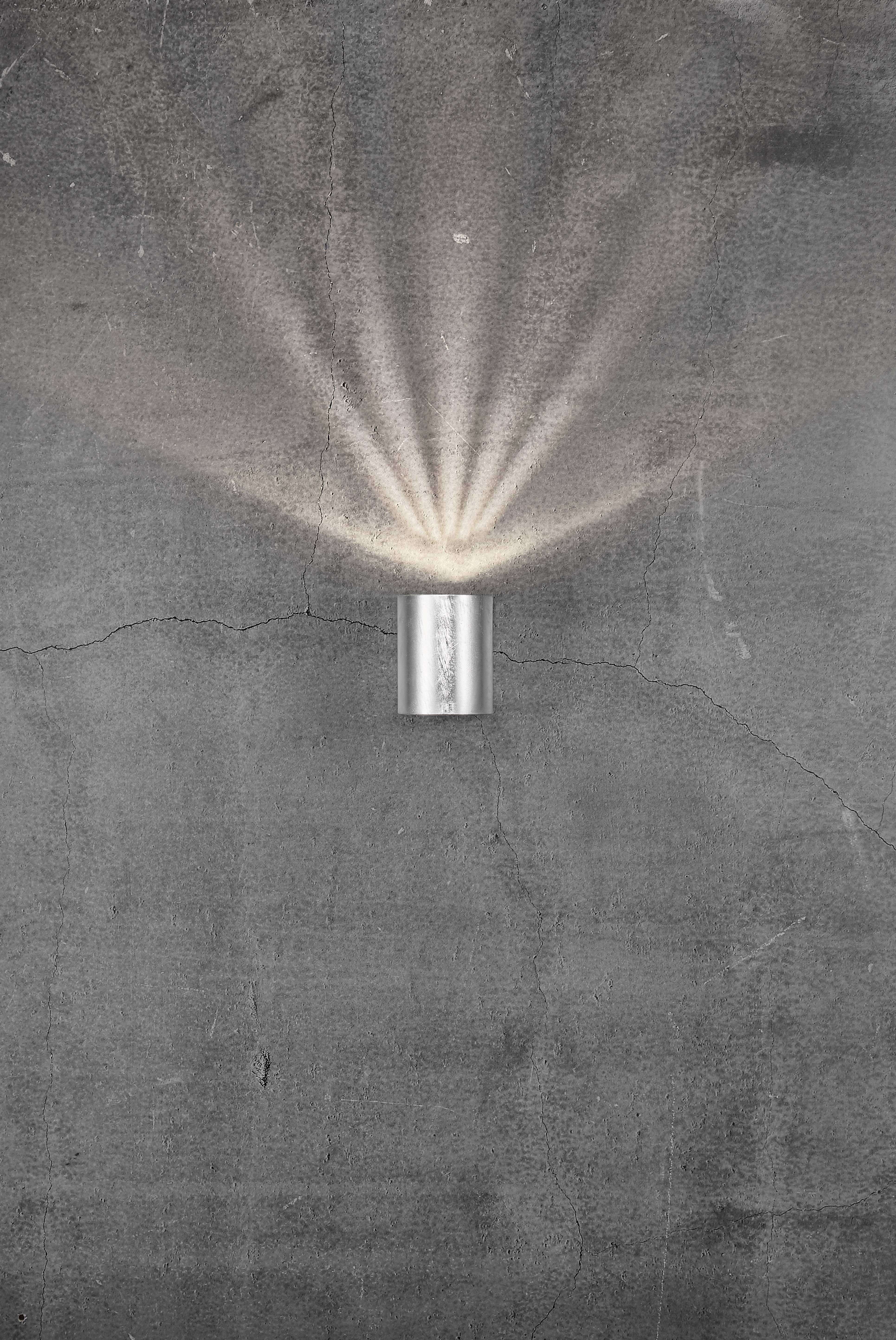 Un applique métallique projette des rayons lumineux en éventail vers le haut et vers le bas sur un mur en béton gris, légèrement fissuré.