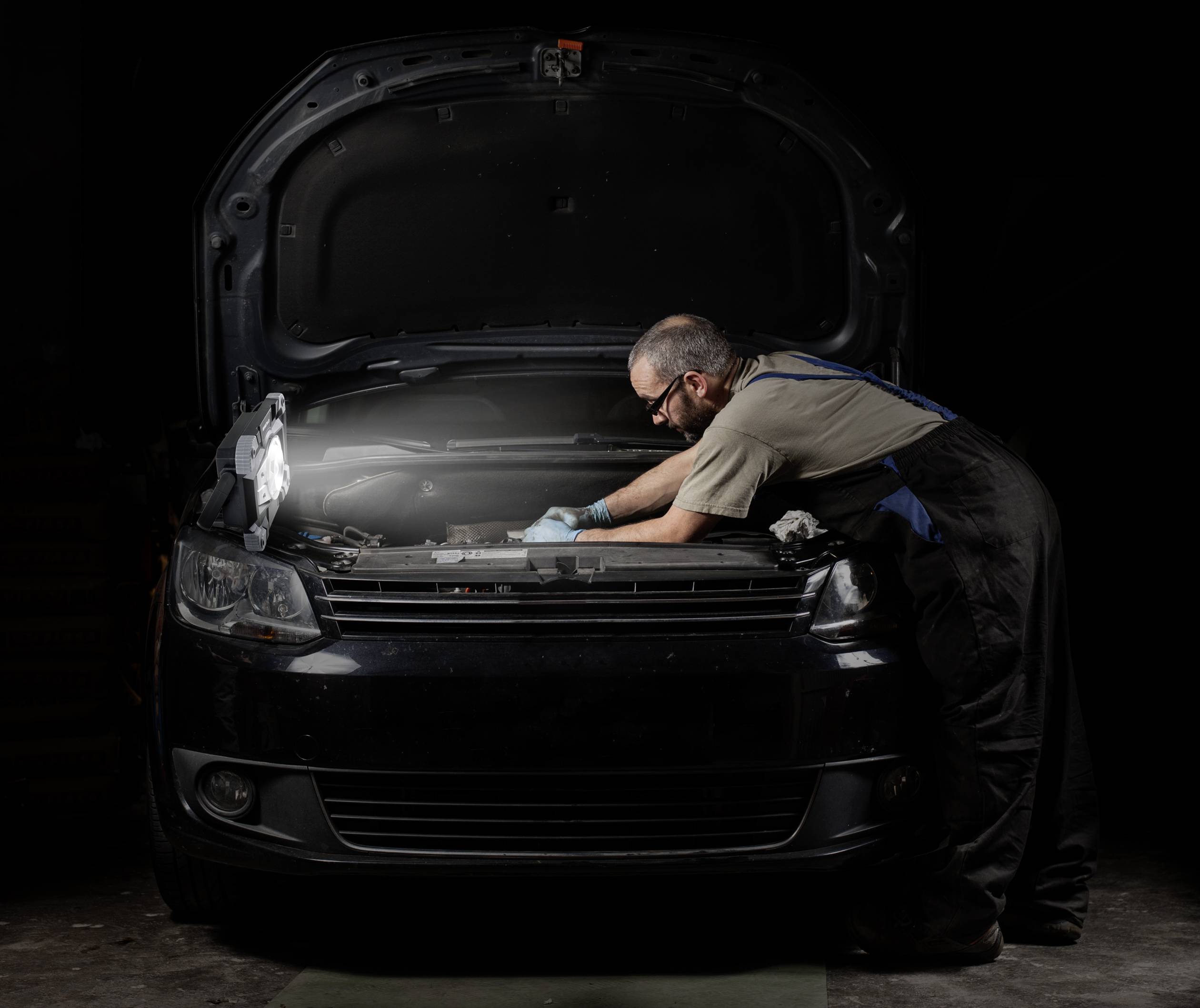Un mécanicien dans un atelier répare une voiture avec le capot ouvert. Il porte des lunettes de protection et des gants, et travaille avec concentration sur le moteur.