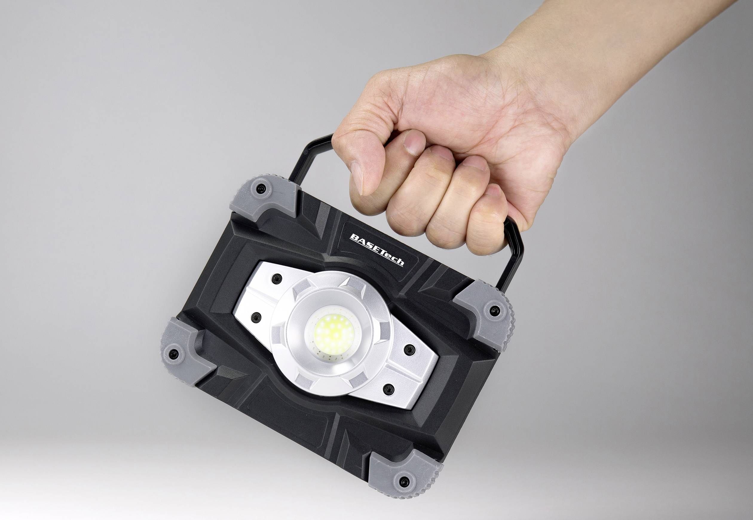 Un projecteur LED est tenu par une main sur une poignée. Le projecteur présente un design robuste, noir et argenté.