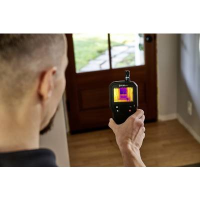 Une personne utilise une caméra thermique dans un couloir pour mesurer les différences de température au niveau d'une porte. L'écran affiche des images thermiques colorées.
