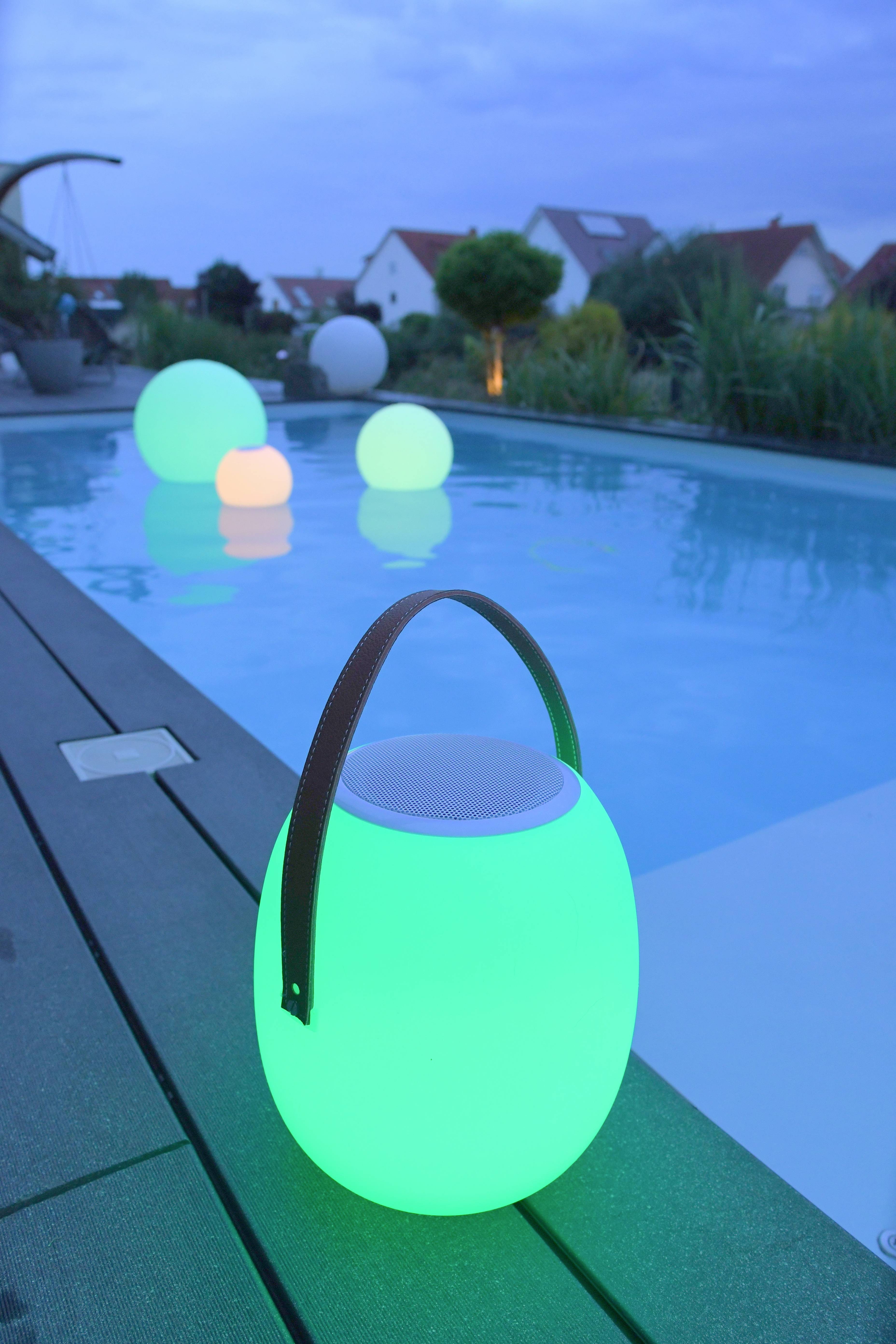 Une piscine au crépuscule avec des lampes sphériques lumineuses bordant les bords et flottant dans l'eau. L'arrière-plan révèle des maisons et des plantes.