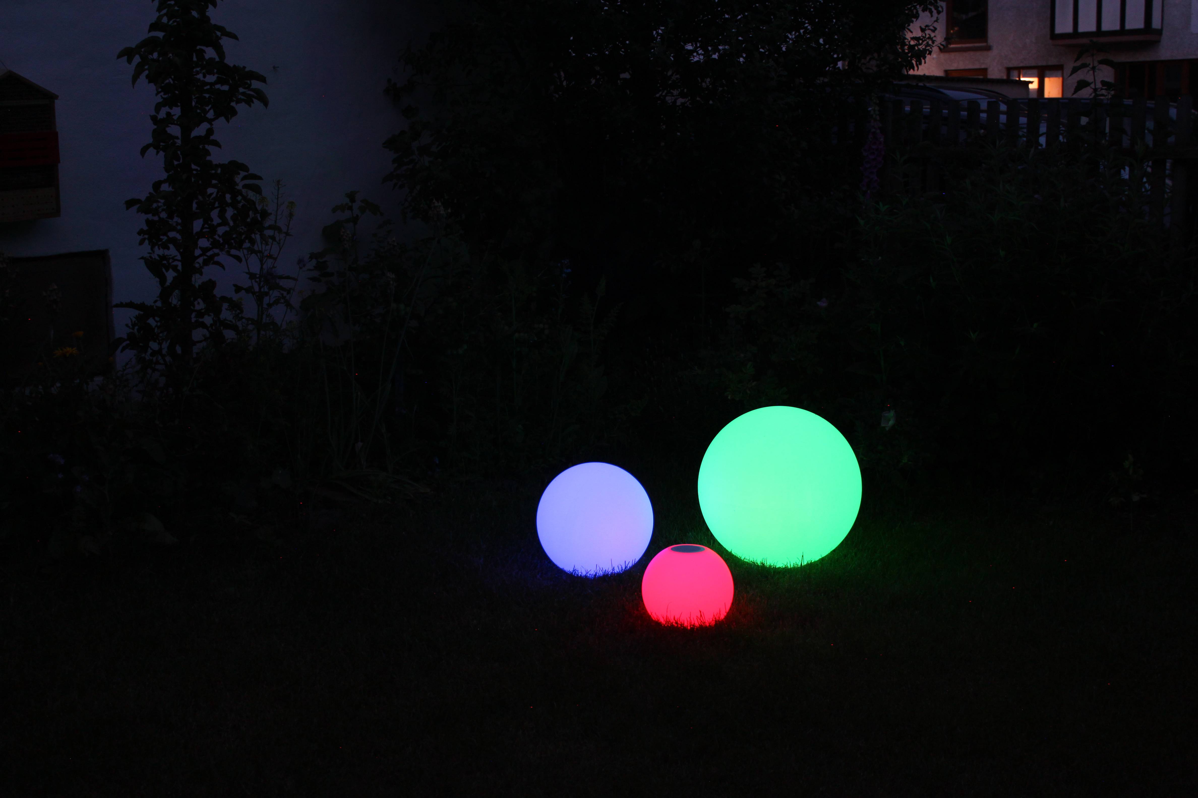 Trois boules lumineuses dans un jardin nocturne : une grande boule verte, une boule bleue moyenne et une petite boule rouge, illuminant l'espace extérieur.