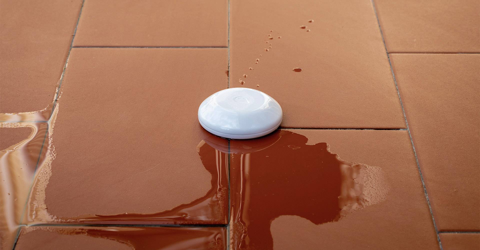Un capteur blanc est posé sur un sol carrelé humide et brun, susceptible de déclencher une alarme eau.