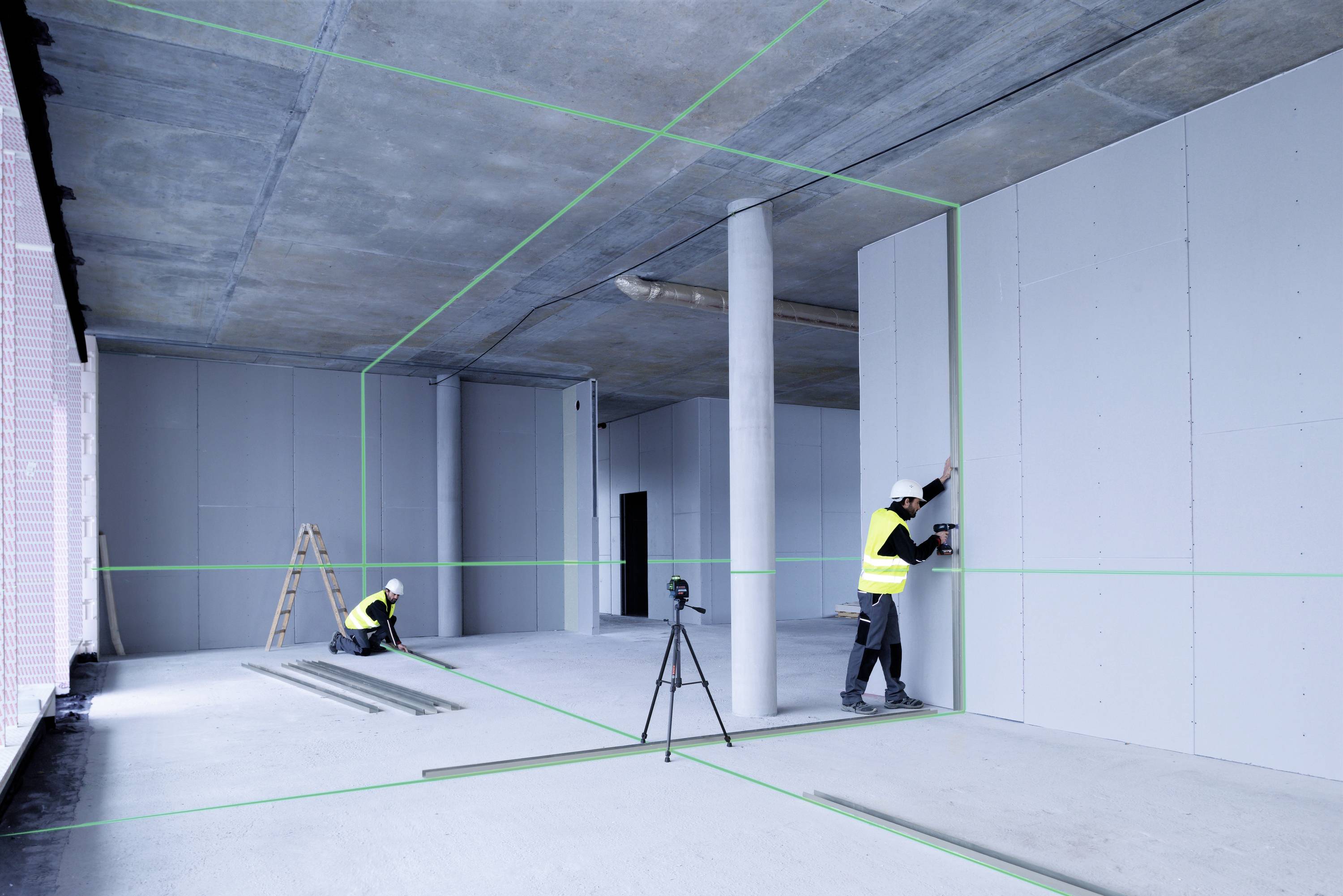 Deux ouvriers du bâtiment installent des cloisons en plaques de plâtre dans un bâtiment. Une ligne laser verte indique l'alignement. Un trépied est placé au centre.