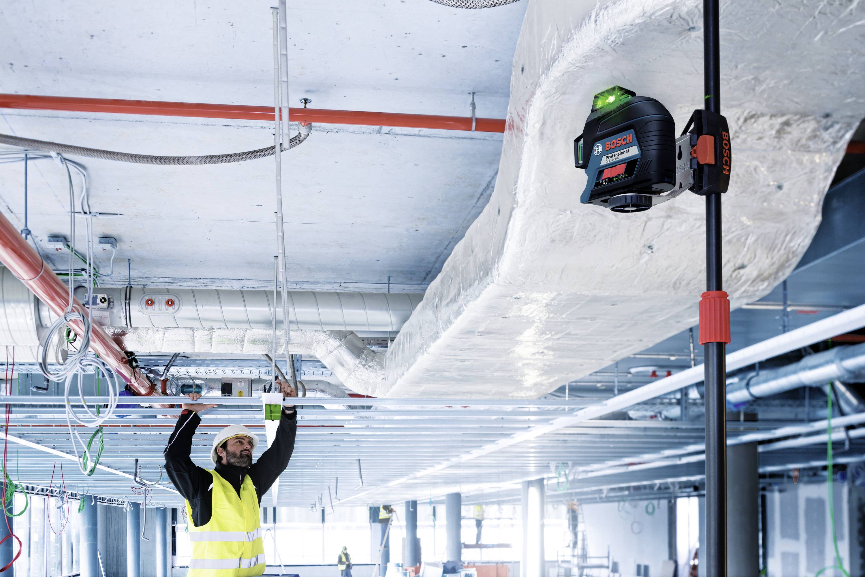 Un ouvrier portant un gilet de sécurité mesure la hauteur du plafond à l'aide d'un outil laser sur un chantier de construction.