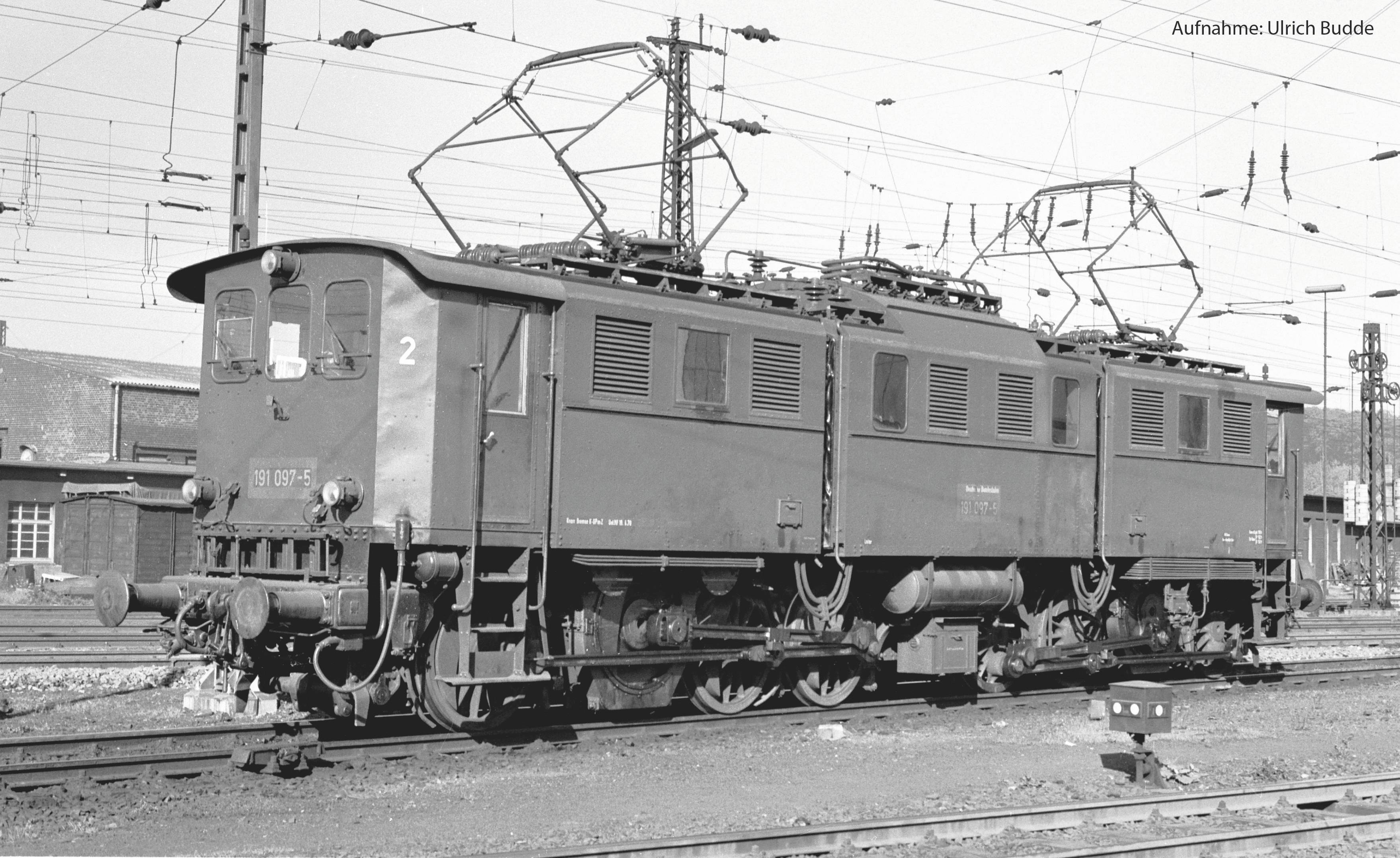 Une photographie historique montre une ancienne locomotive électrique sur une voie ferrée. Deux parties de la locomotive sont connectées entre elles.