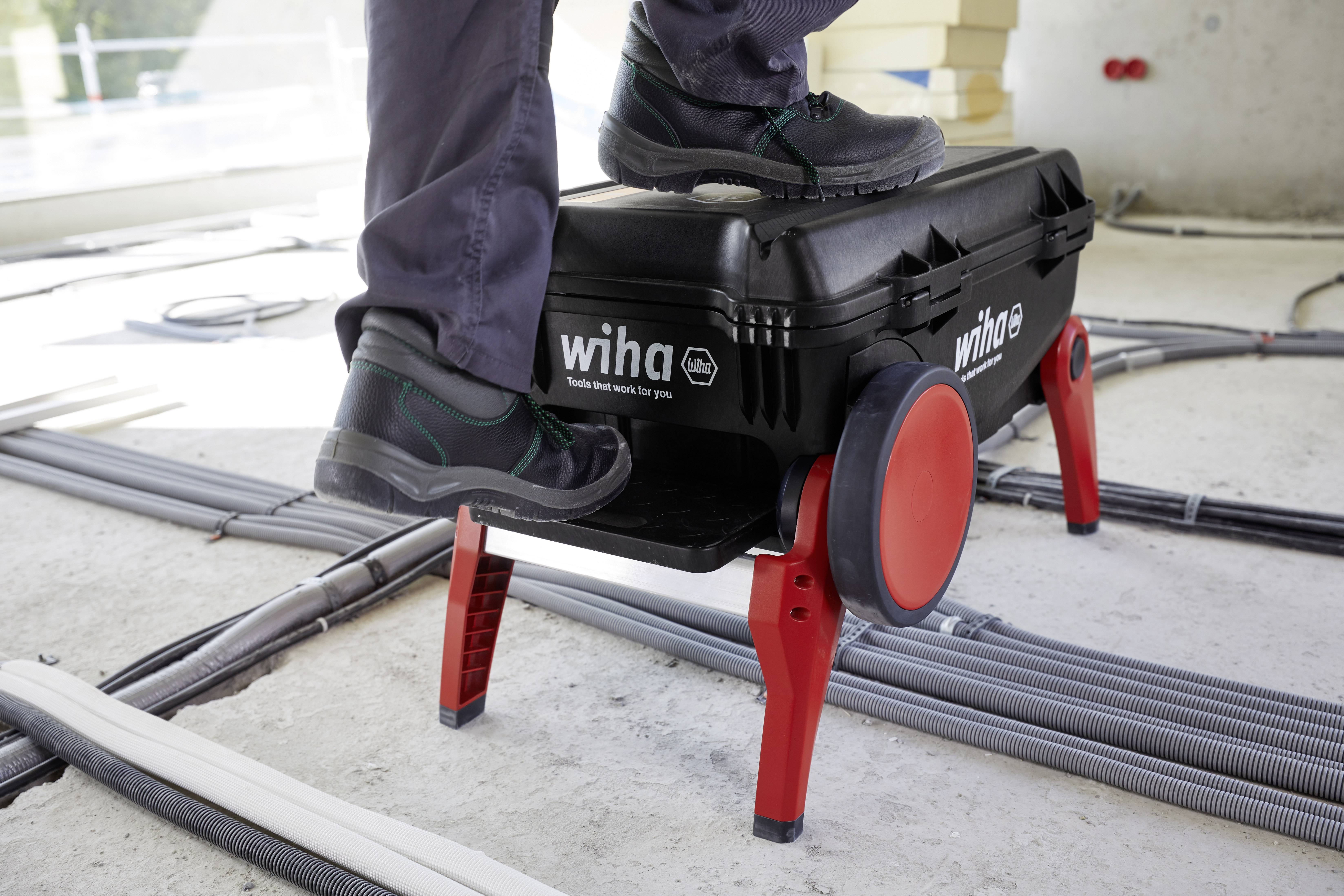 Une personne monte sur une boîte à outils noire avec des pieds rouges, posée sur un sol en câbles. La marque 'wiha' est visible.