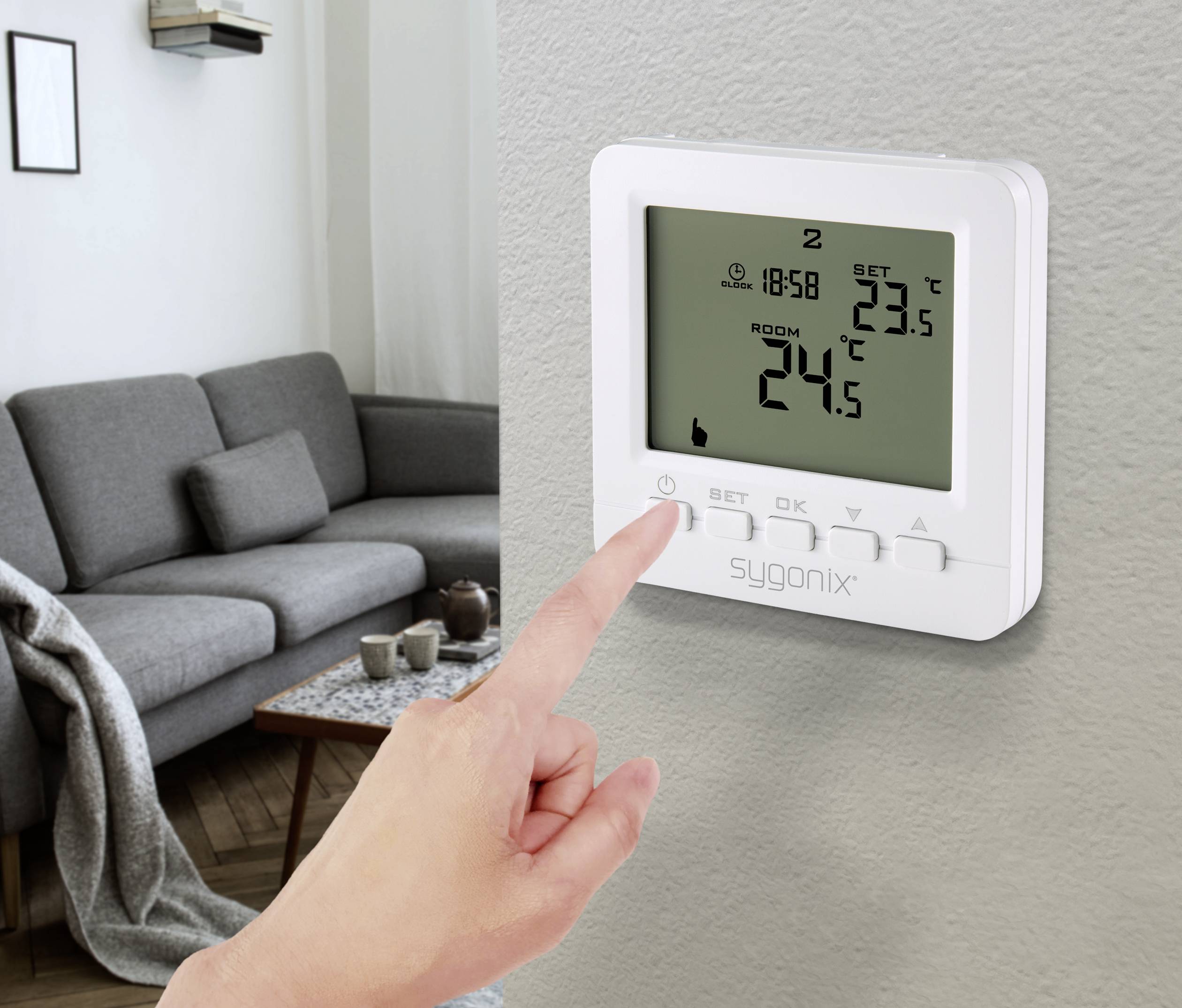 Une personne règle un thermostat numérique à 24 degrés Celsius. Un salon avec un canapé est visible en arrière-plan.