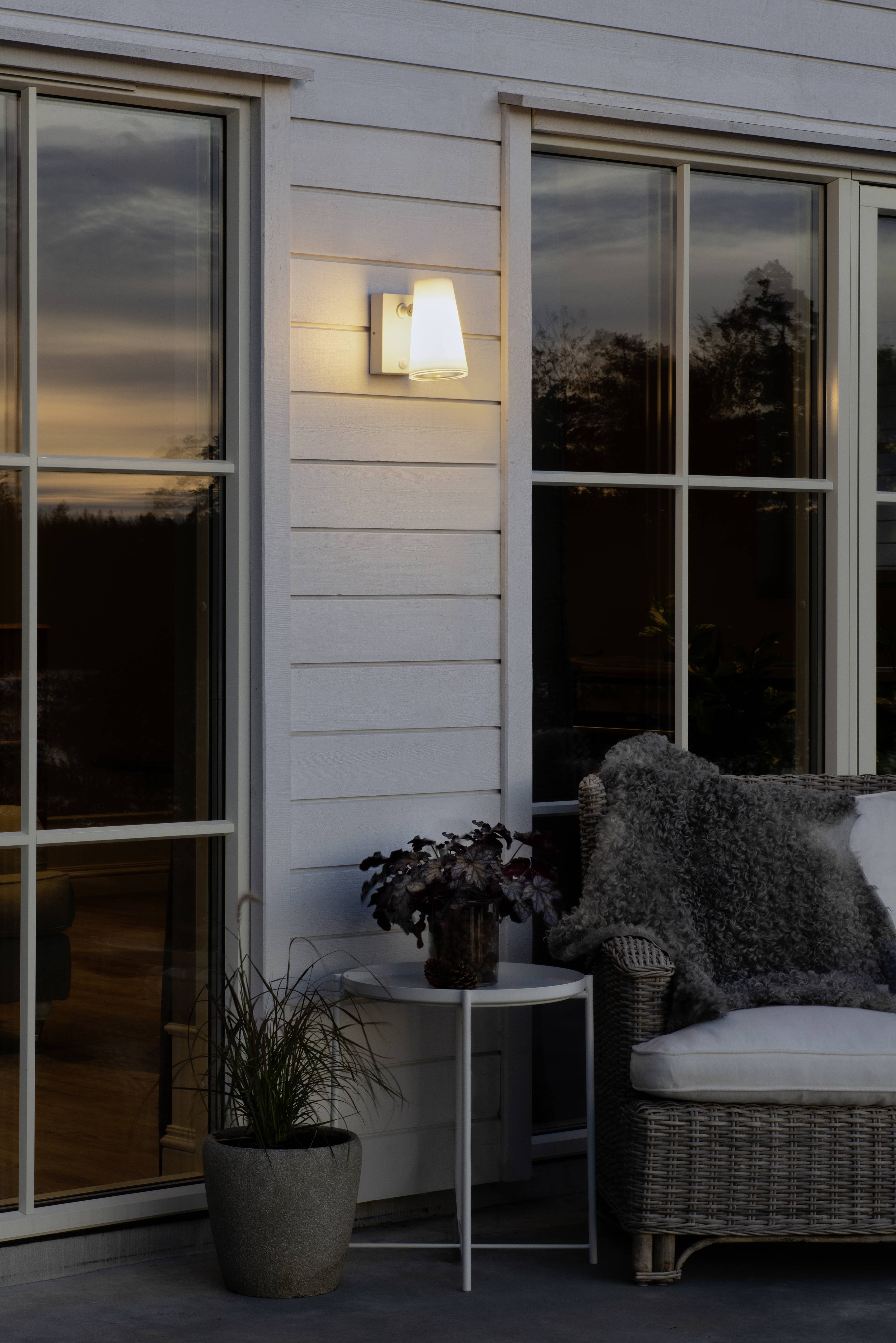 Une terrasse au coucher du soleil révèle des meubles en rotin confortables avec un jeté en fourrure douce à côté d'un applique murale illuminée.