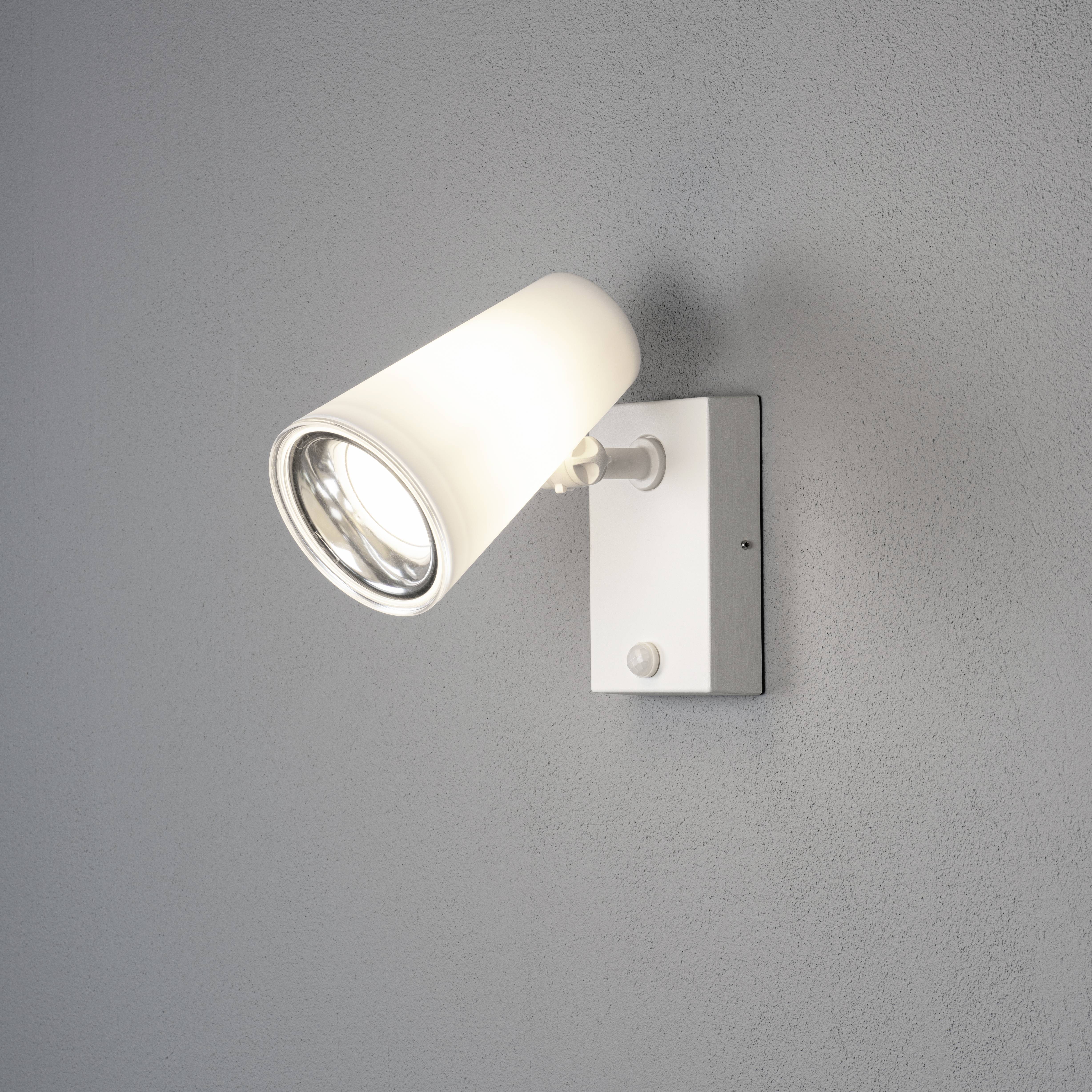 Un luminaire mural moderne blanc avec un abat-jour cylindrique fixé à un mur gris, éclairant vers la gauche.