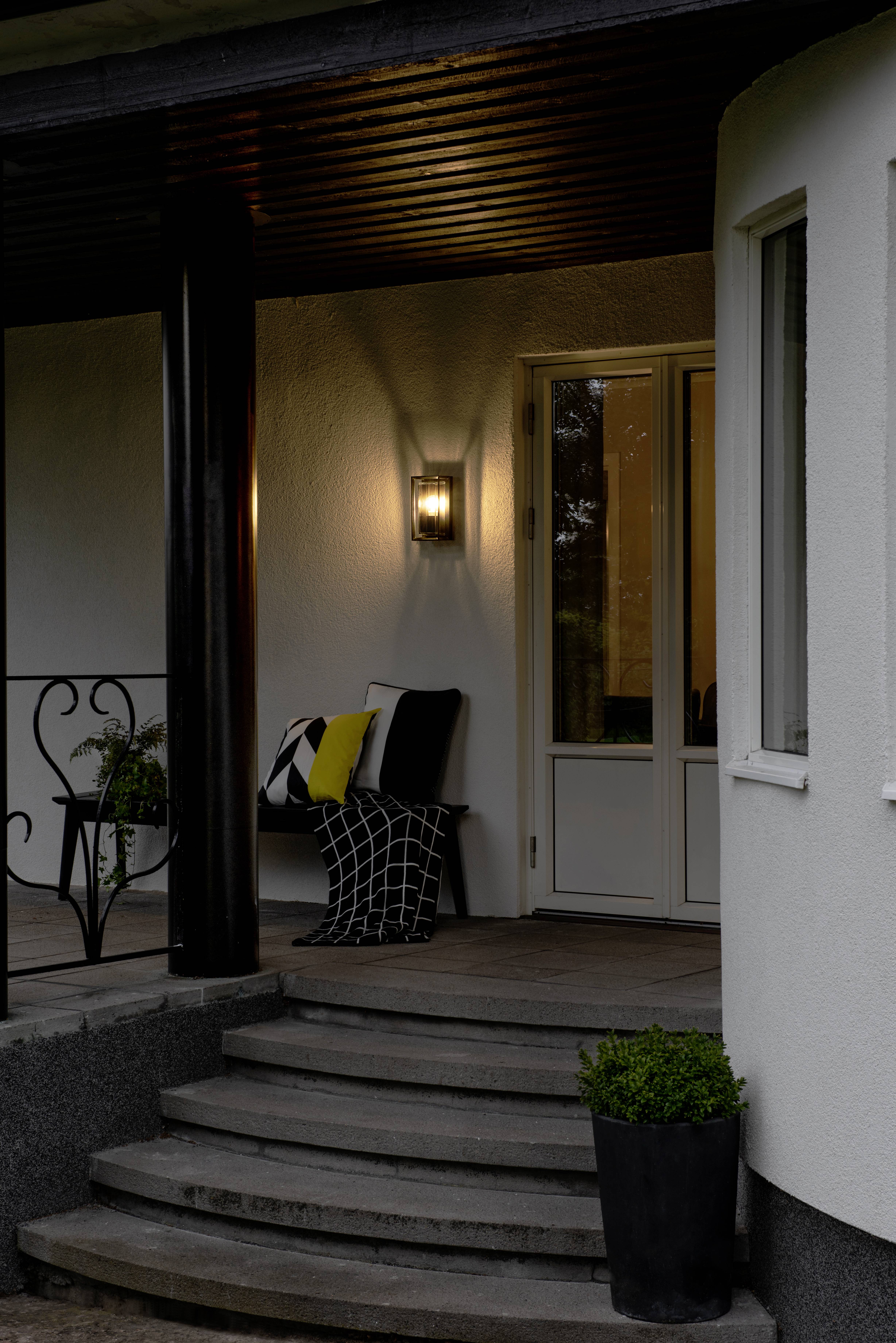 Une entrée de maison en soirée avec une lampe allumée, des rideaux ouverts, des marches et une plante devant la porte.