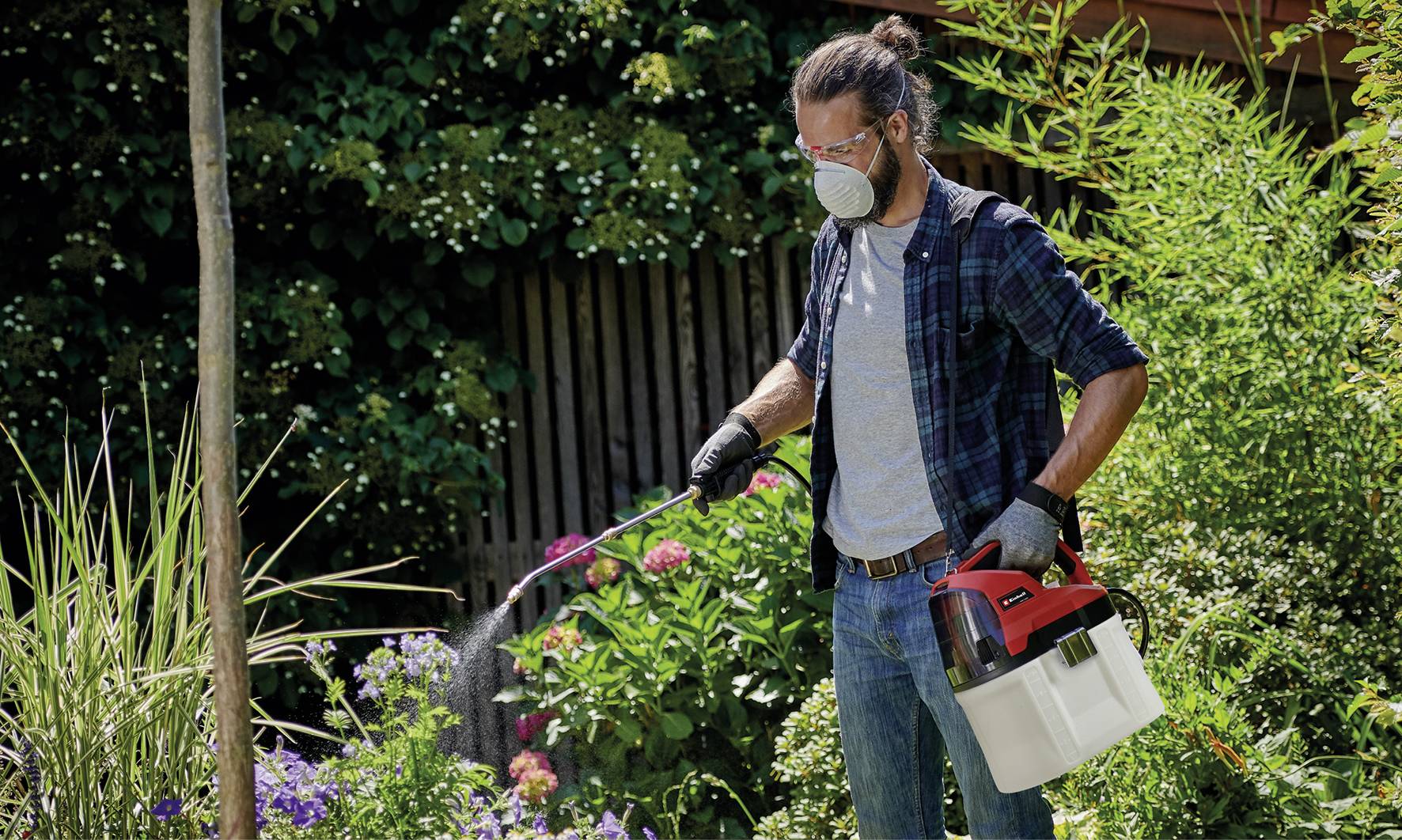 Une personne dans le jardin pulvérise des plantes avec un pulvérisateur de jardin. Elle porte un masque de protection, des gants et des lunettes. Elle est entourée de buissons et de fleurs.