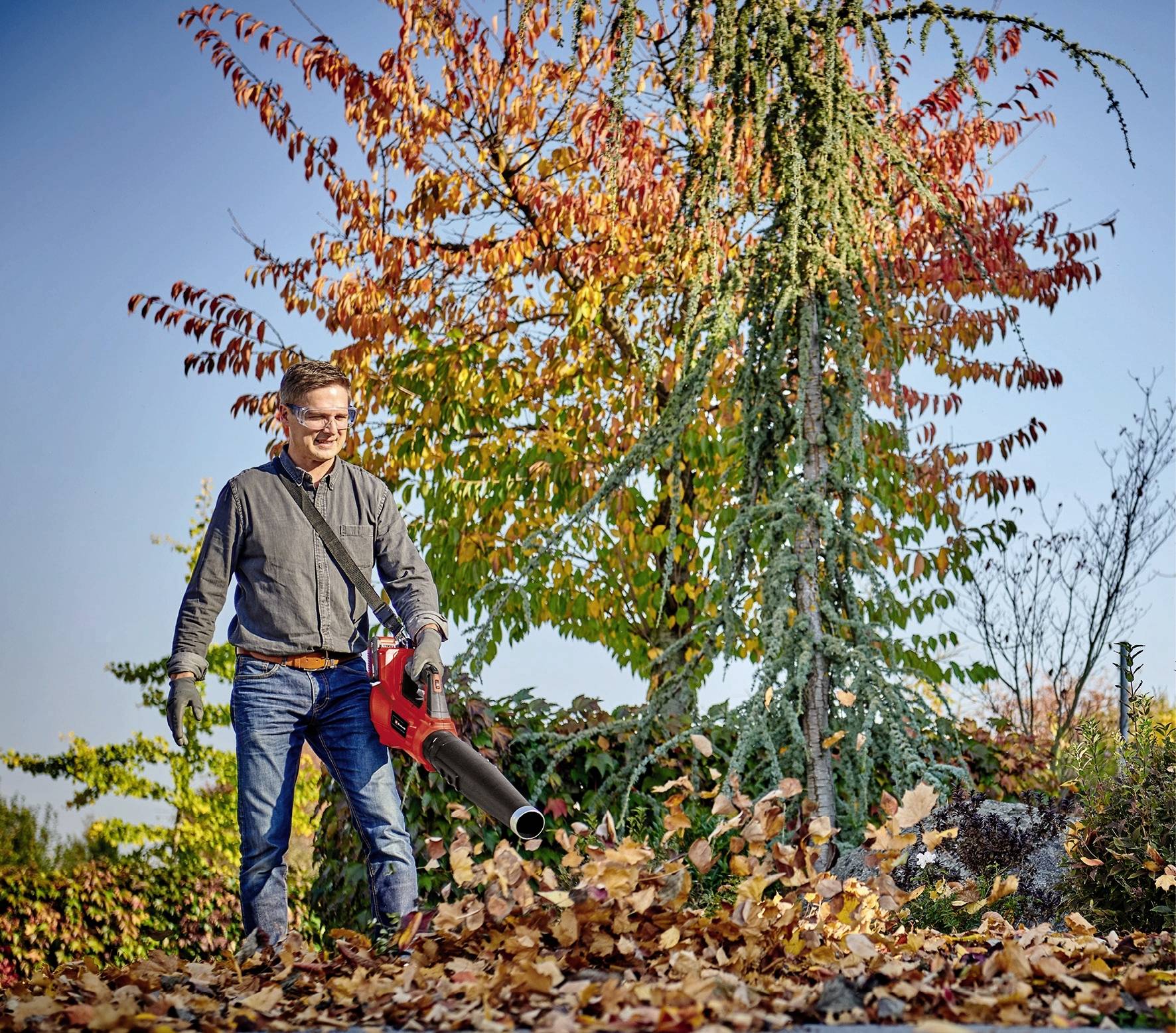 Un homme utilise un souffleur de feuilles pour disperser les feuilles d'automne dans un jardin. En arrière-plan, on peut voir des arbres aux couleurs automnales.