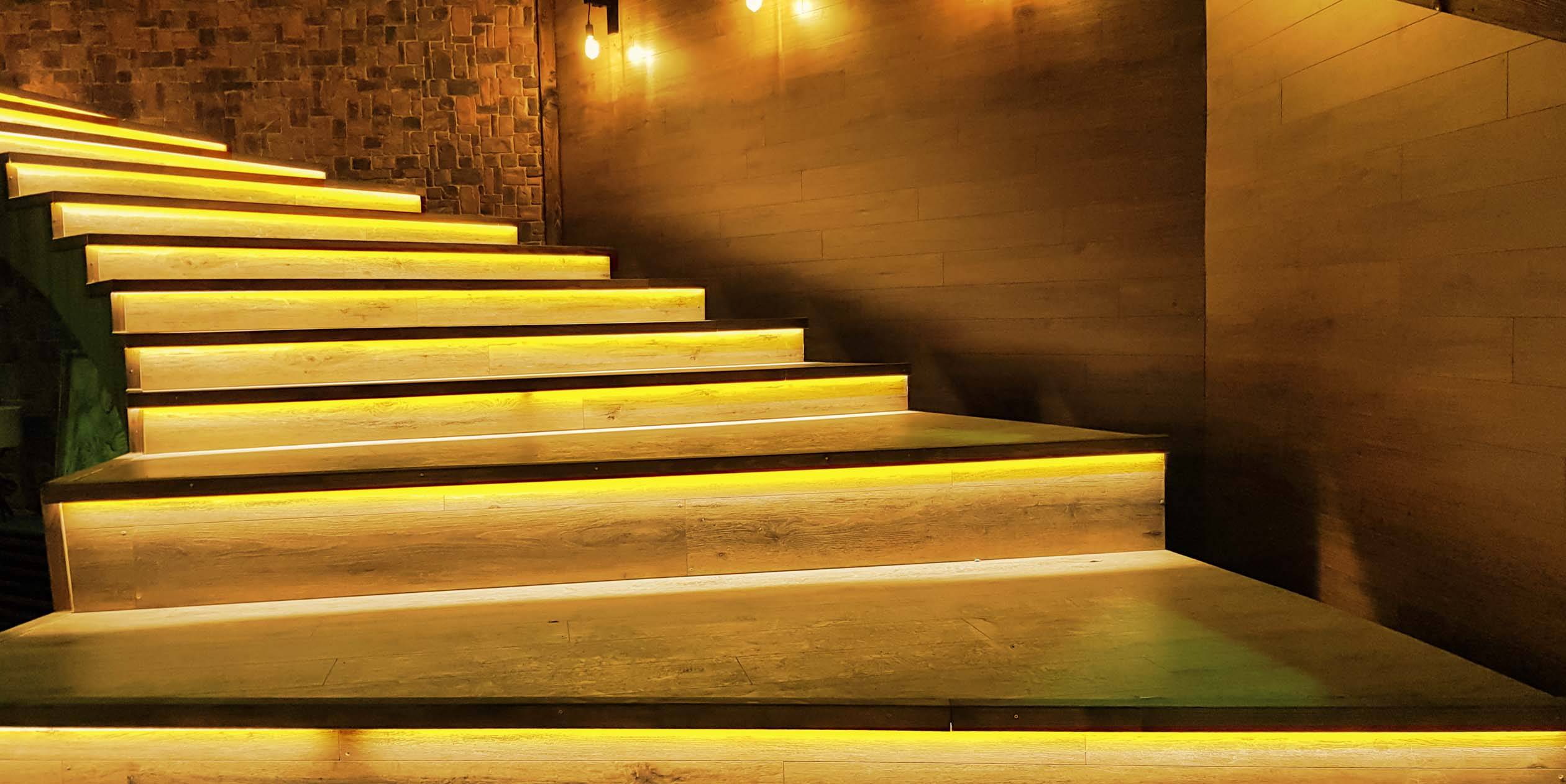 Escalier baigné d'une lumière chaleureuse qui met en valeur les marches, dans une pièce aux murs de briques et de bois faiblement éclairée.