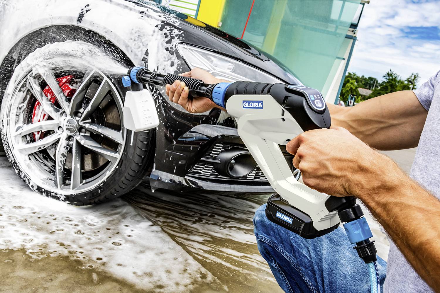 'Une personne lave une voiture avec un nettoyeur haute pression, pulvérise de l'eau sur la roue arrière et de la mousse recouvre la voiture.'