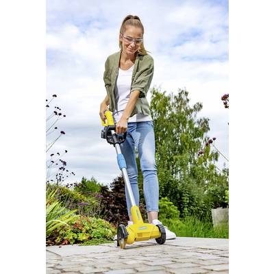 Personne portant des lunettes de protection utilisant un appareil de jardinage jaune sur un chemin pavé, entourée de plantes. Ciel clair en arrière-plan.