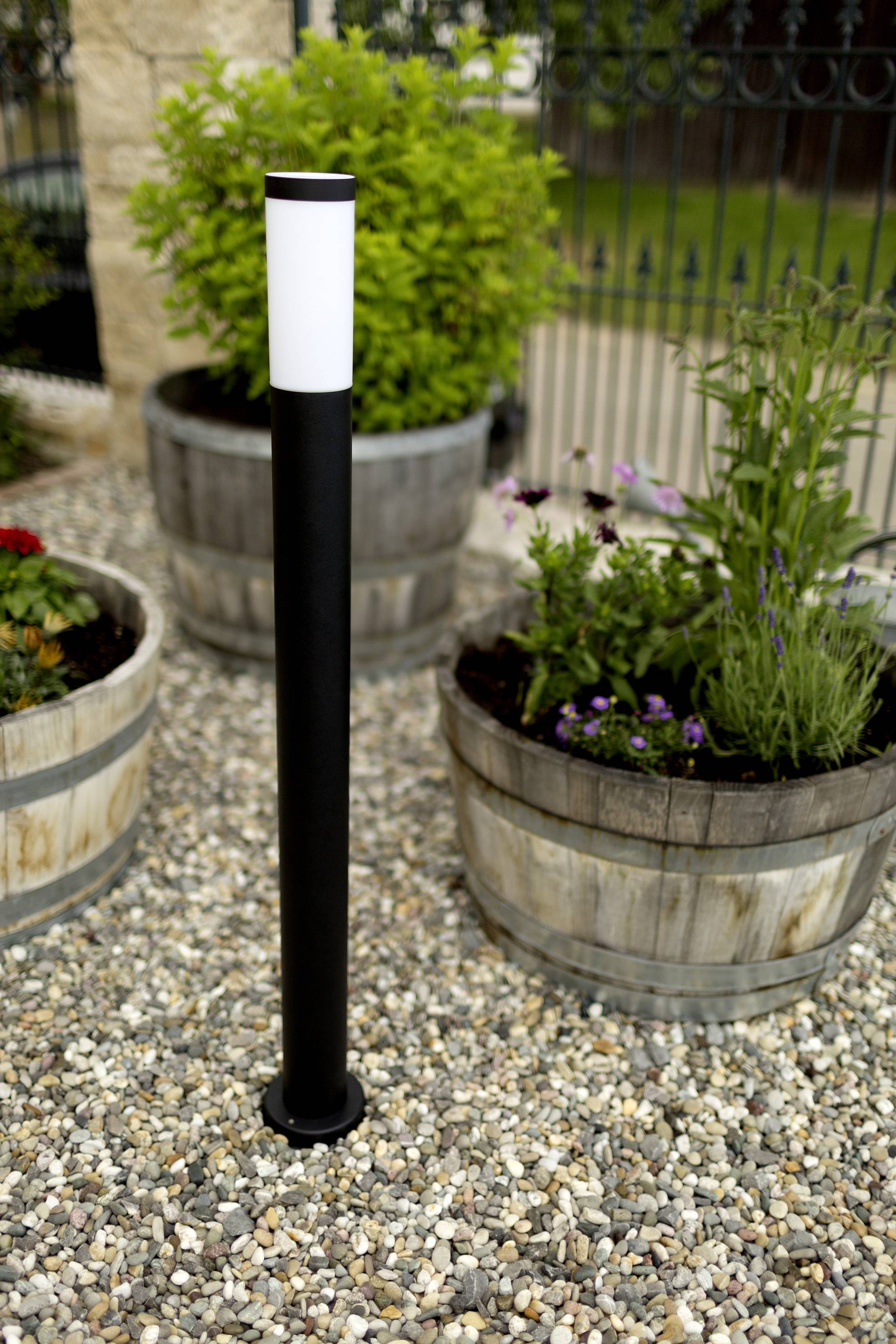 Lampadaire noir avec une tête cylindrique blanche, situé sur un chemin de gravier, flanqué de bacs en bois contenant des plantes vertes et fleuries.