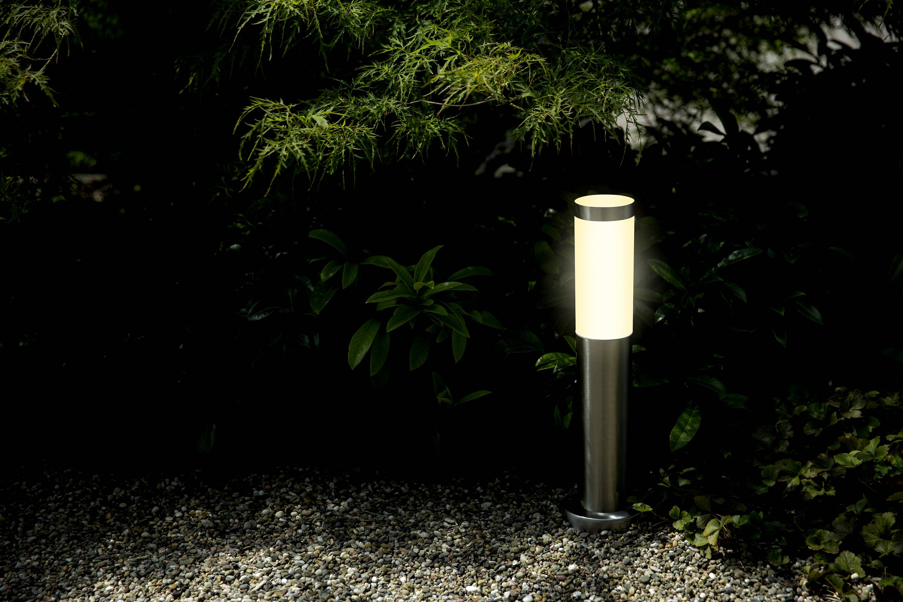 Une lampe de jardin illuminée se dresse dans un lit de graviers, entourée de plantes vertes dans l'obscurité.