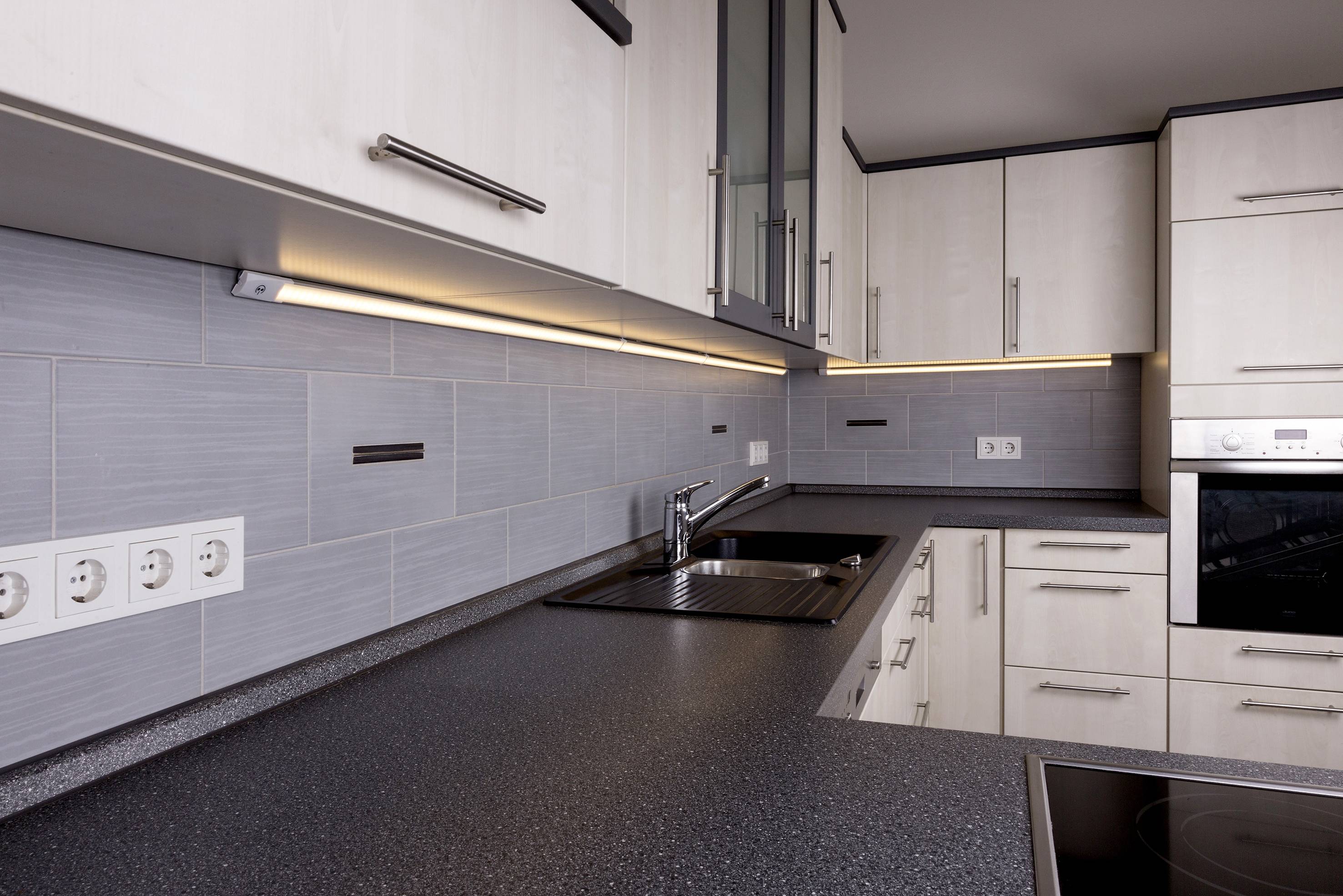 Cuisine moderne avec évier d'angle, plan de travail gris et éclairage sous-jacent. Des meubles hauts blancs et une plaque de cuisson intégrée sont visibles.