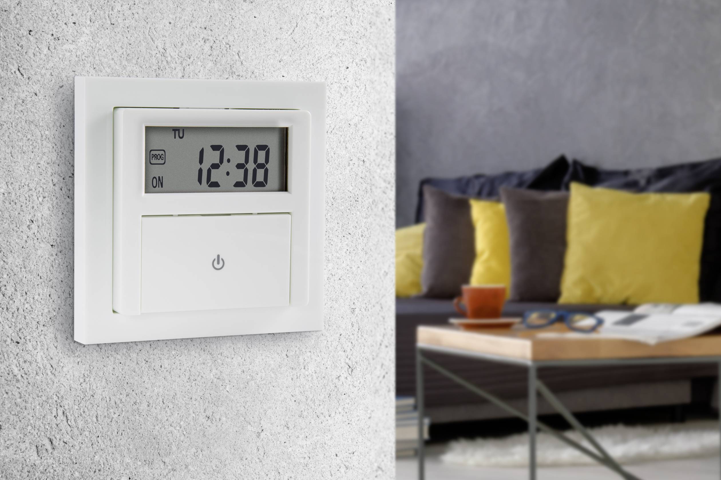 Une horloge programmable murale affiche '12:38'. En arrière-plan, on aperçoit un salon moderne avec un canapé et une table.