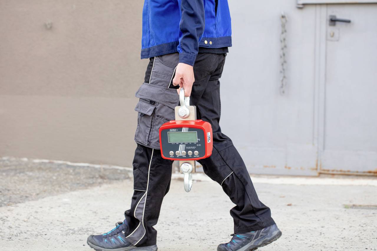 Une personne en tenue de travail porte un appareil portable rouge avec un écran, probablement destiné à des mesures industrielles, sur un sol en béton.