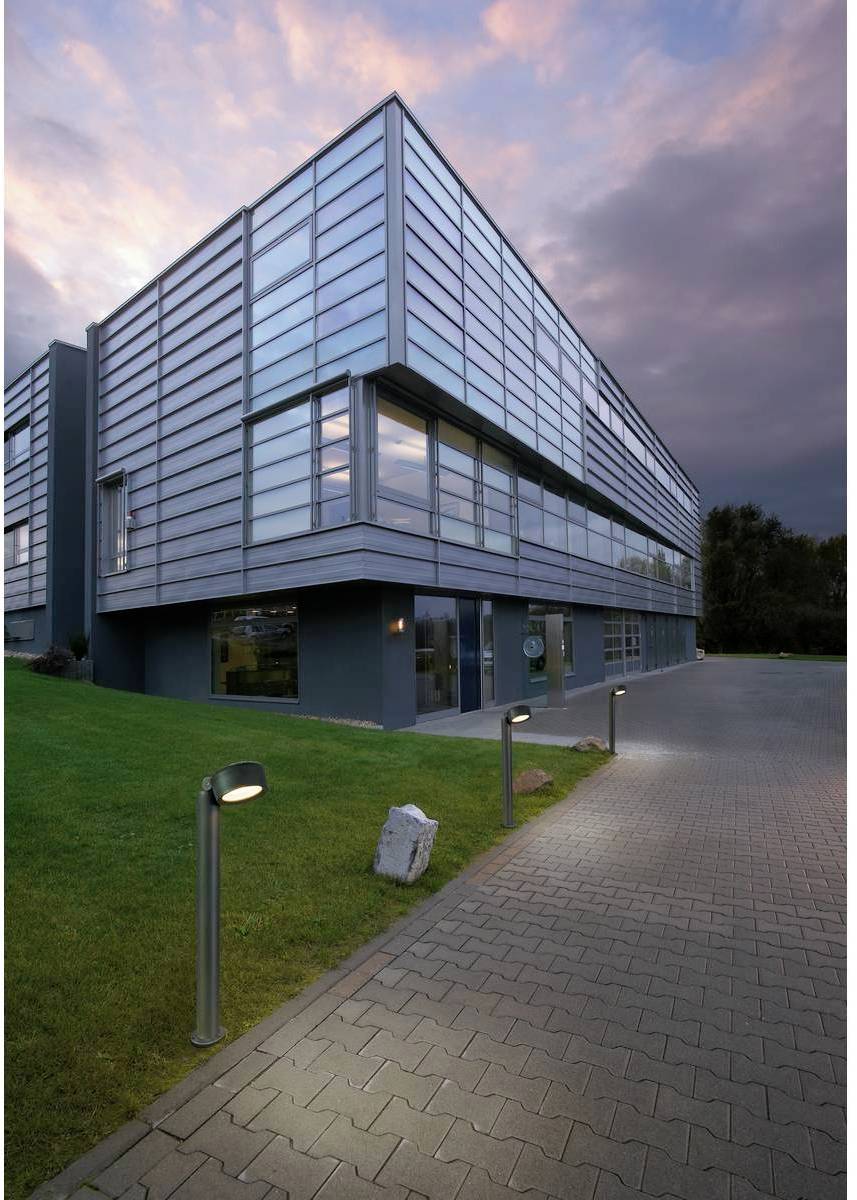 Un immeuble de bureaux moderne à façade vitrée au crépuscule. Le ciel est nuageux avec des tons roses et gris. Un chemin pavé mène au bâtiment.
