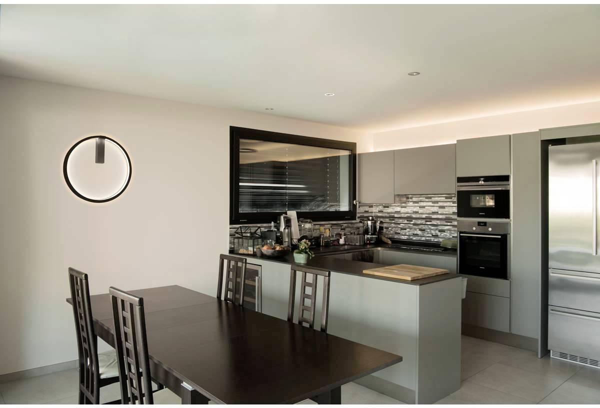 Cuisine moderne avec espace repas, table en bois, cinq chaises, placards gris, appareils en acier inoxydable, crédence carrelée et lampe ronde.