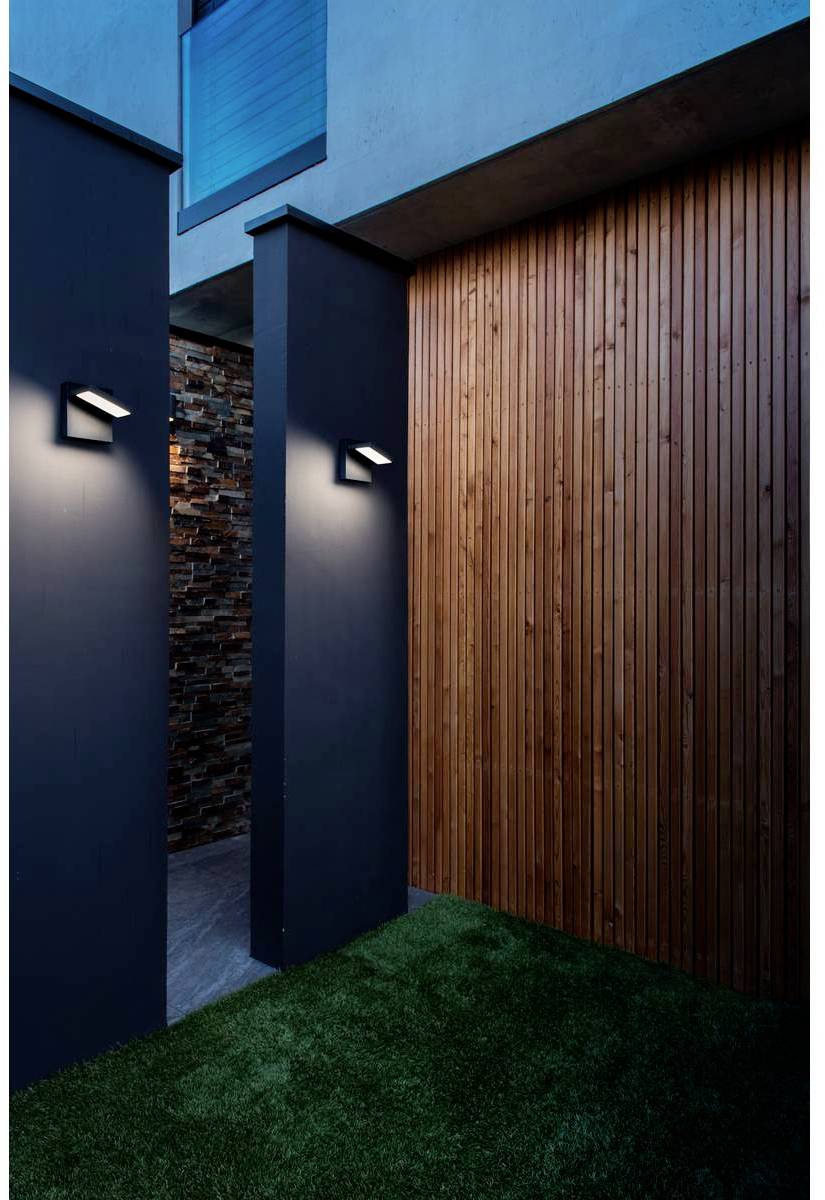 Espace extérieur moderne éclairé, avec revêtement en bois, appliques murales et gazon synthétique, situé entre deux murs de bâtiments.