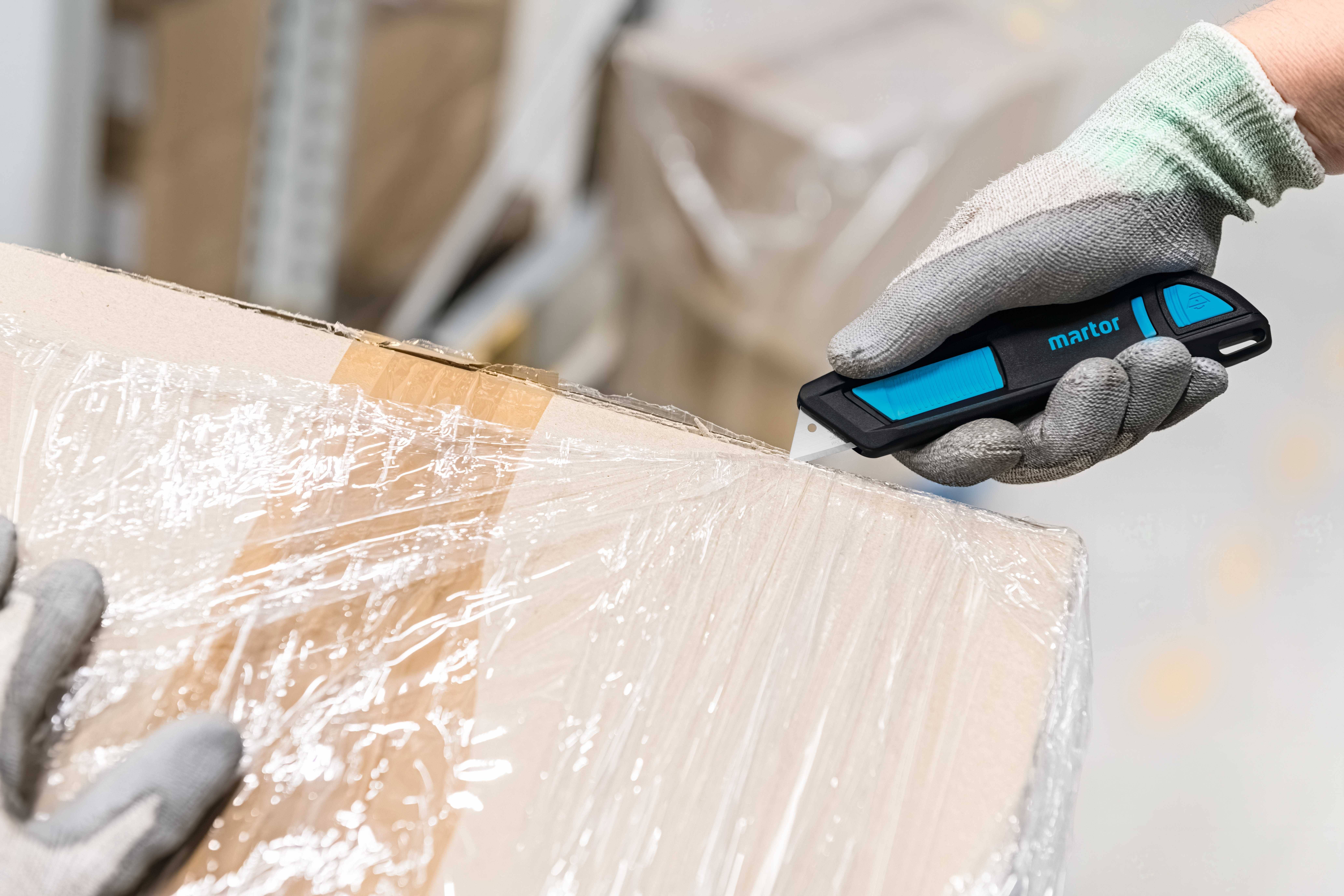 Une personne portant des gants ouvre prudemment un carton en coupant un film de protection avec un cutter de sécurité bleu.