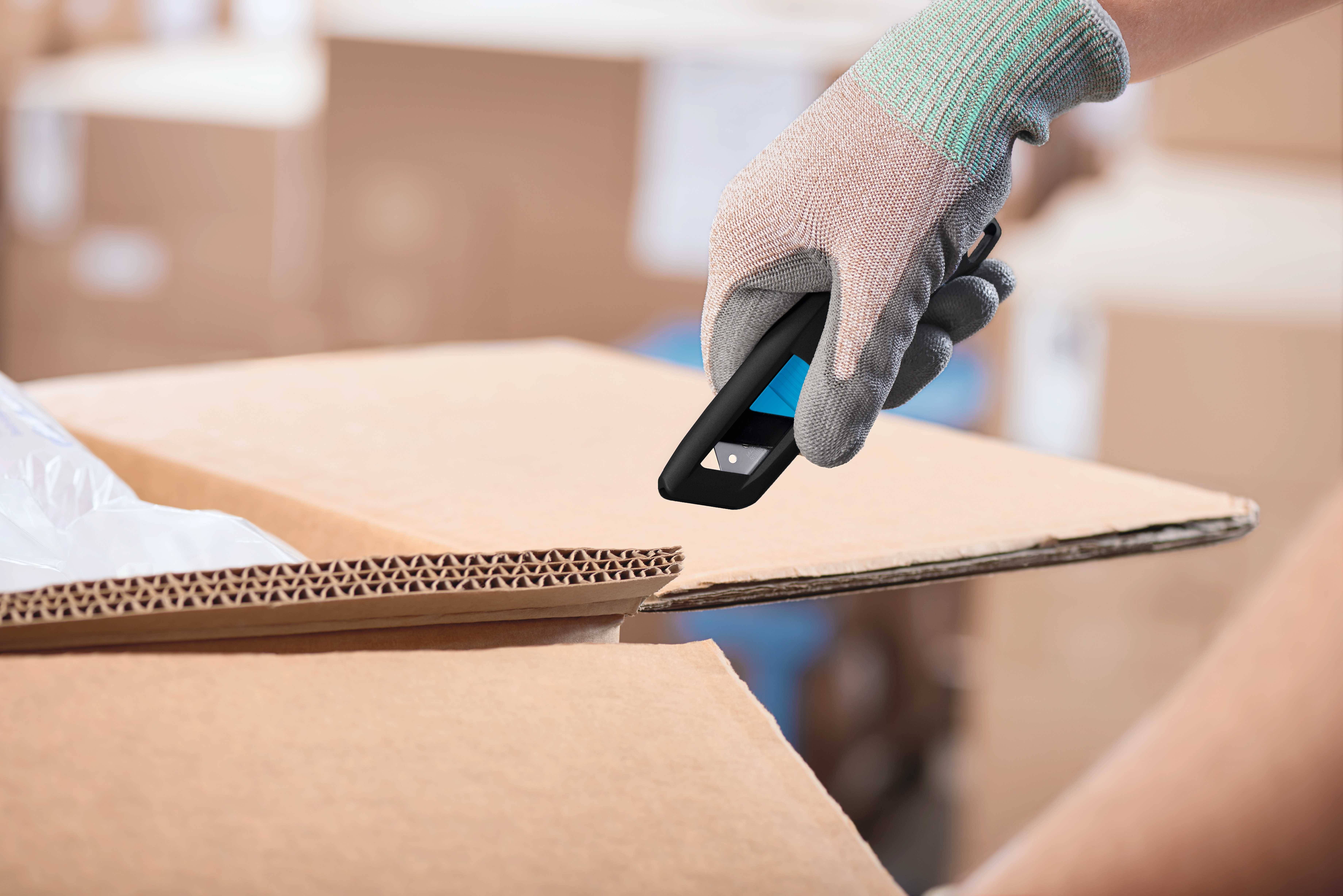Une main gantée ouvre délicatement un carton avec un cutter. D'autres cartons sont visibles en arrière-plan.