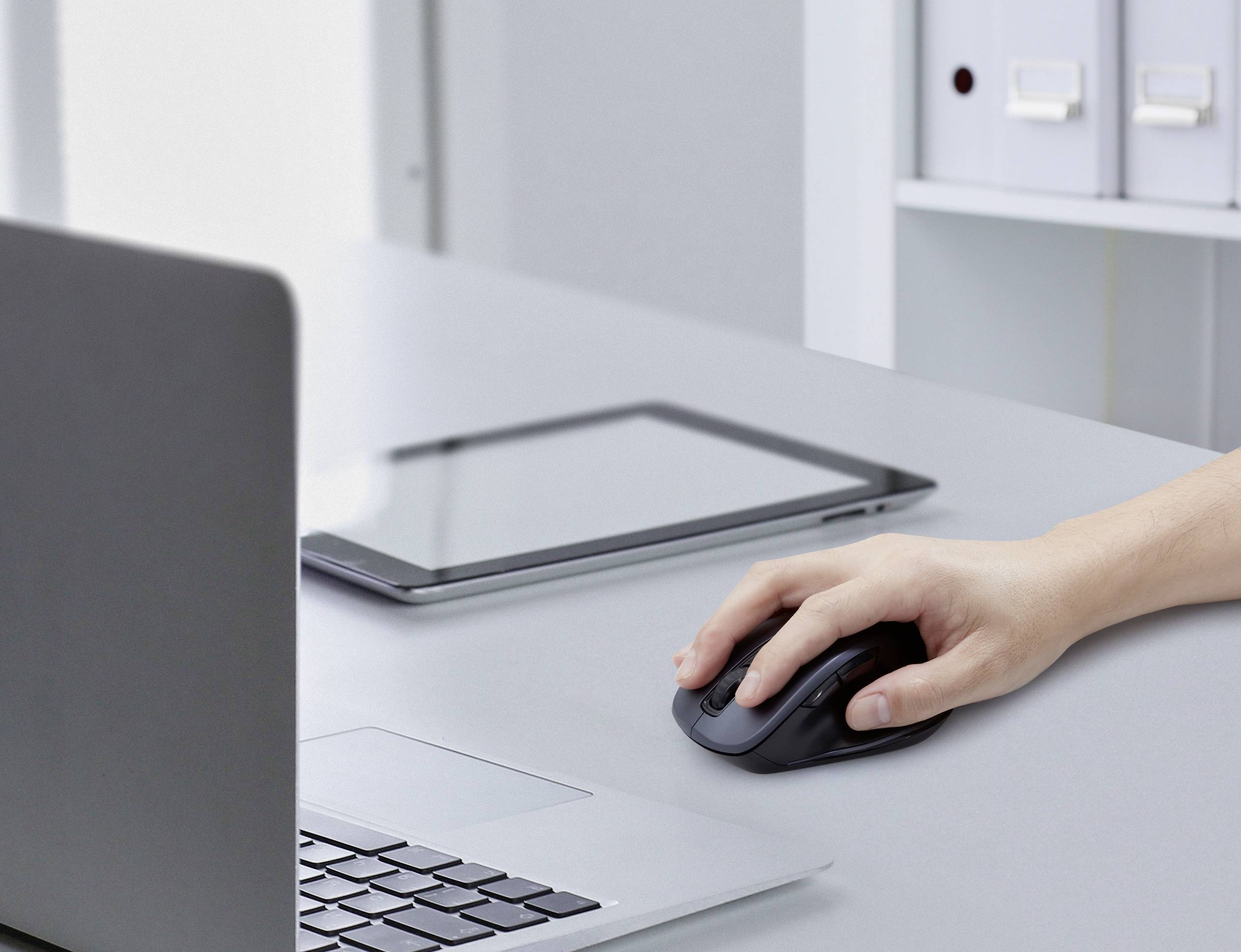 Une main contrôle une souris sans fil à côté d'un ordinateur portable et d'une tablette sur un bureau. Des dossiers sont visibles en arrière-plan.