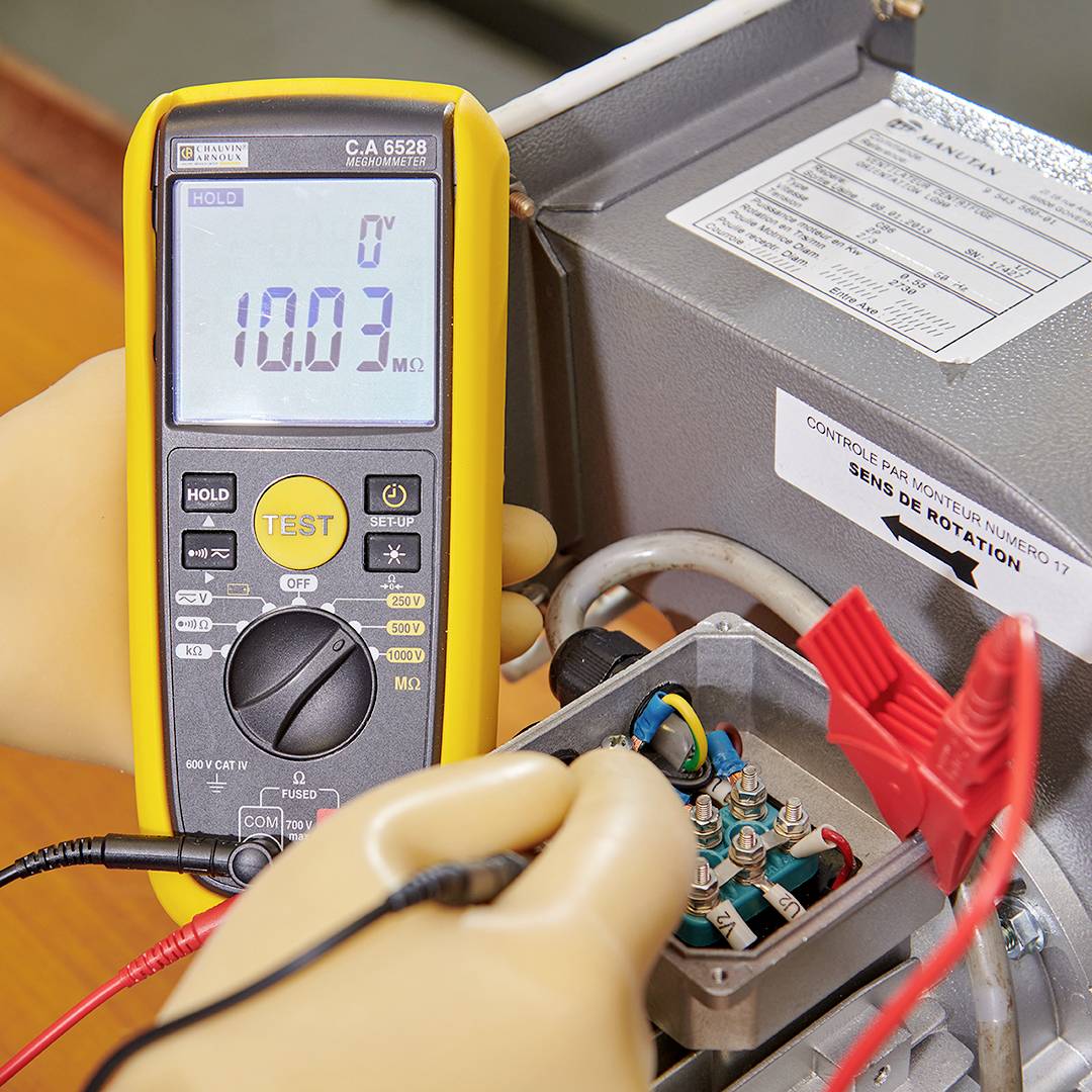 Un multimètre affiche '10,03 MΩ', tenu par une personne portant des gants, mesurant un circuit électrique dans un boîtier métallique.