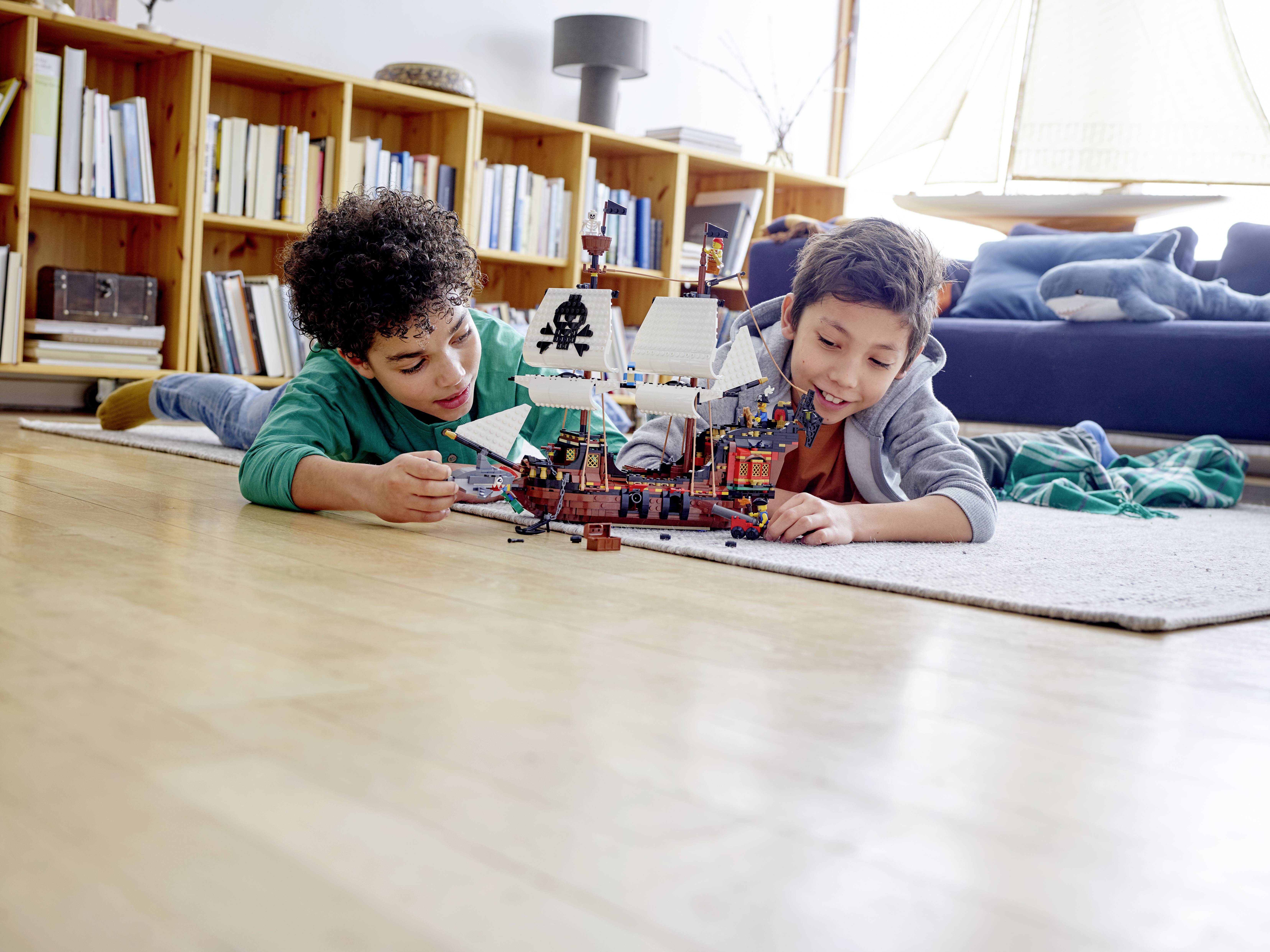 Deux enfants sont allongés sur le tapis et jouent avec un modèle de bateau de pirates. En arrière-plan, on aperçoit une bibliothèque et un canapé.