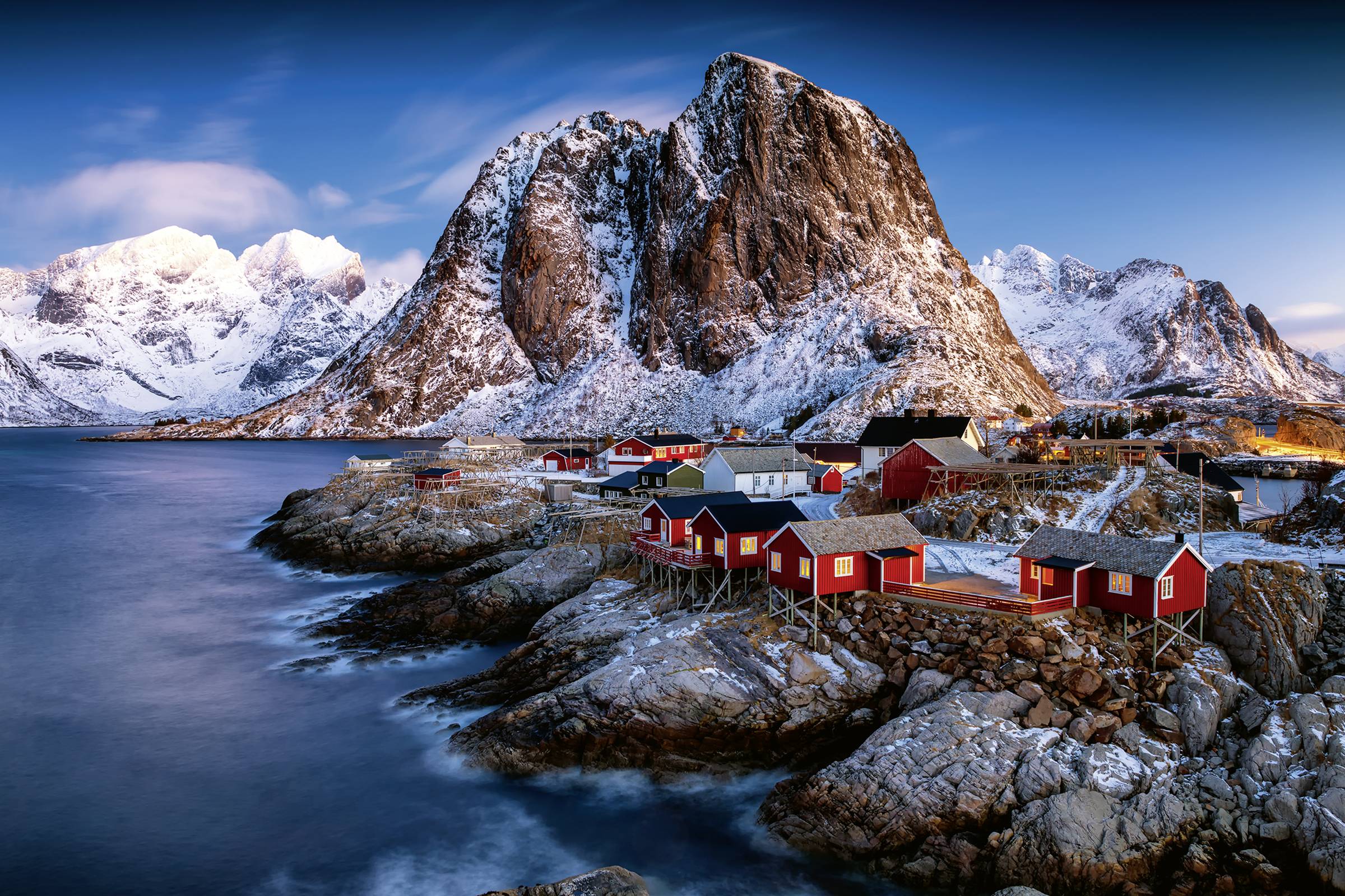 Un paysage côtier pittoresque avec des montagnes enneigées, des maisons en bois rouges sur la rive et une eau calme et bleue au premier plan.