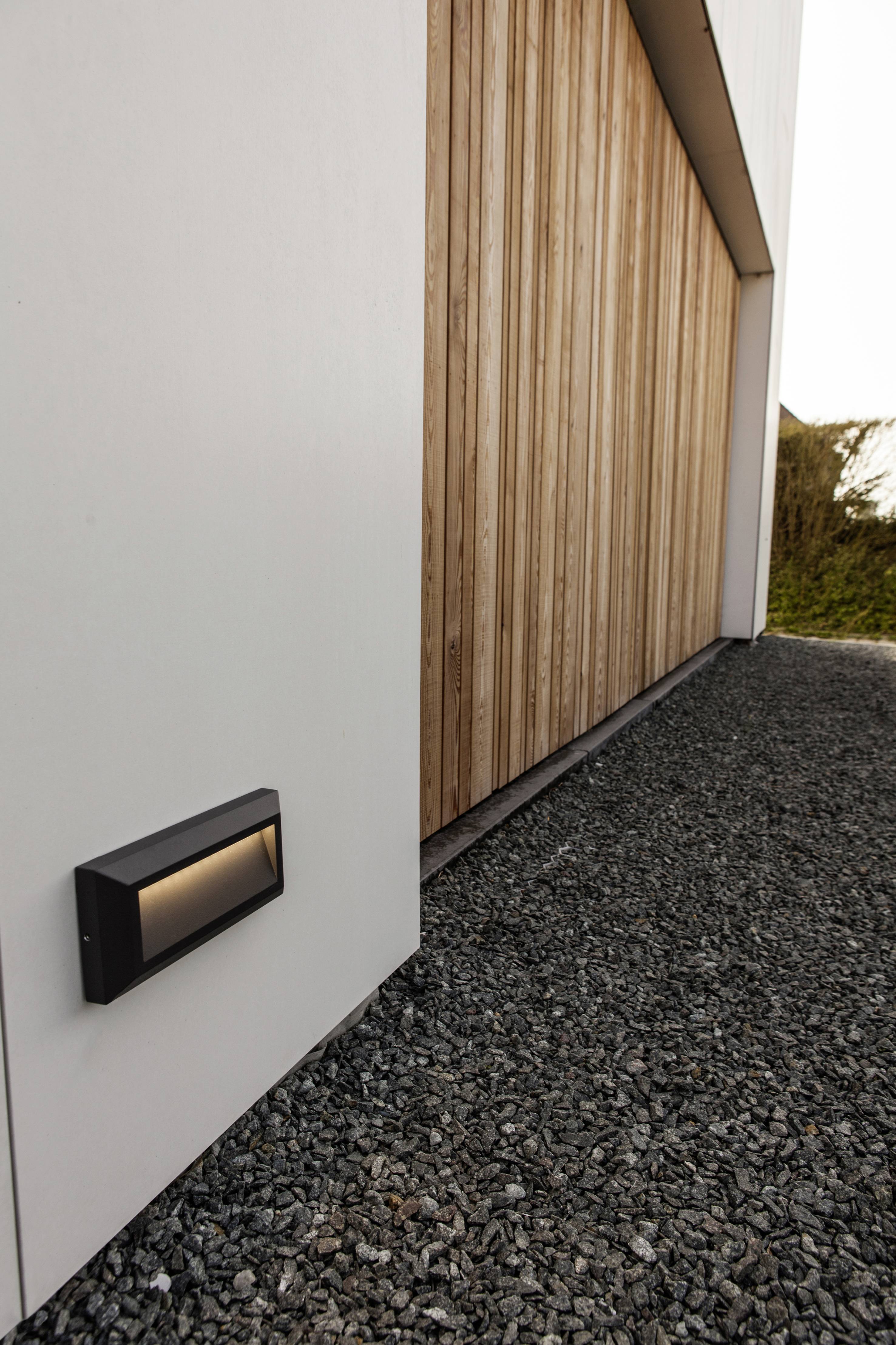 Un garage en bois moderne avec un chemin de gravier. Un luminaire extérieur est installé sur le mur blanc, illuminant le chemin.