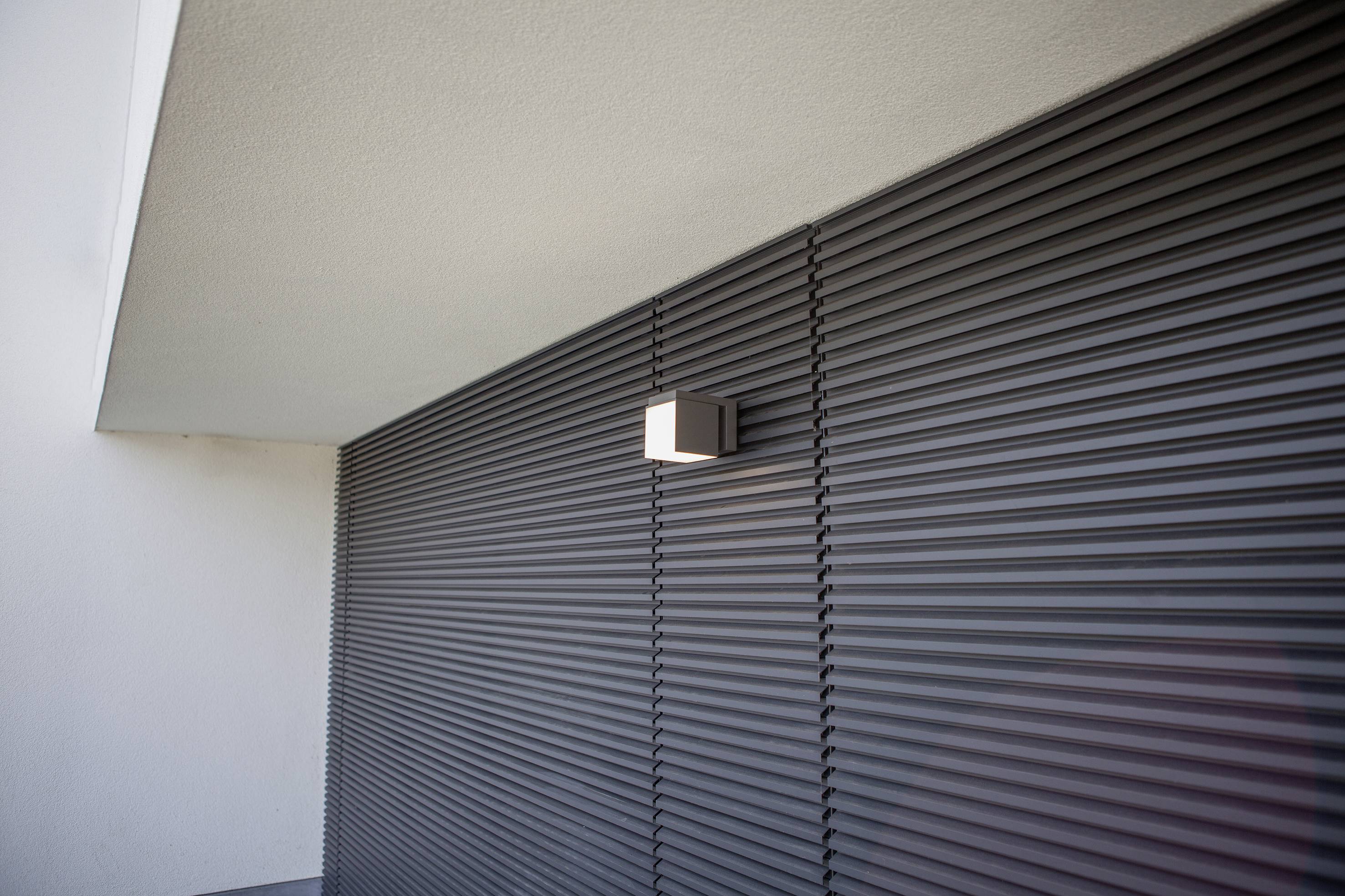Mur extérieur d'un bâtiment avec un revêtement gris moderne et nervuré. Une lampe rectangulaire brille intensément sur le mur, près du toit.
